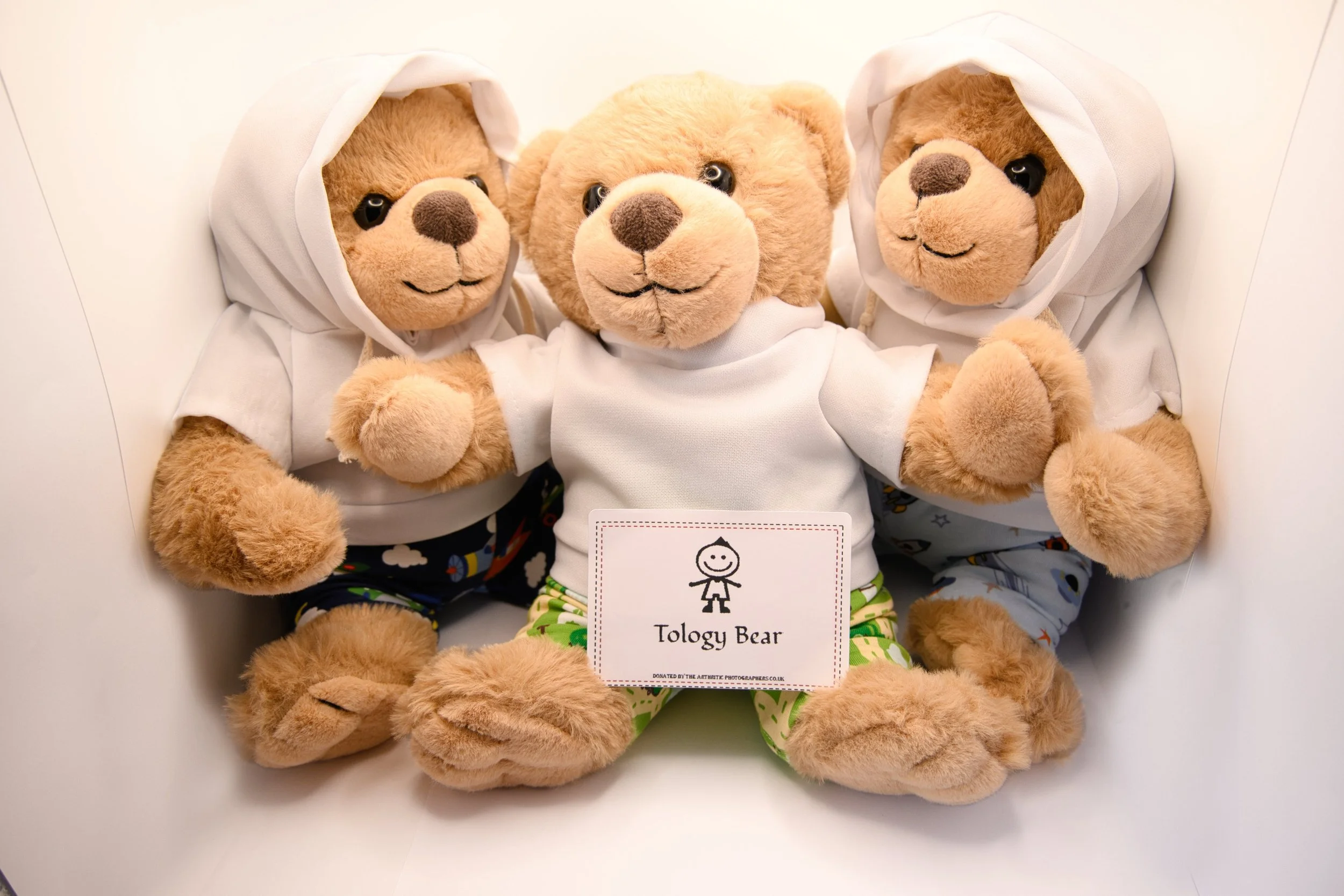 Three plush teddy bears with brown fur, wearing white hoodies and colorful pants, sitting together on a white background. One bear has a tag that says "Tology Bear" attached to it.