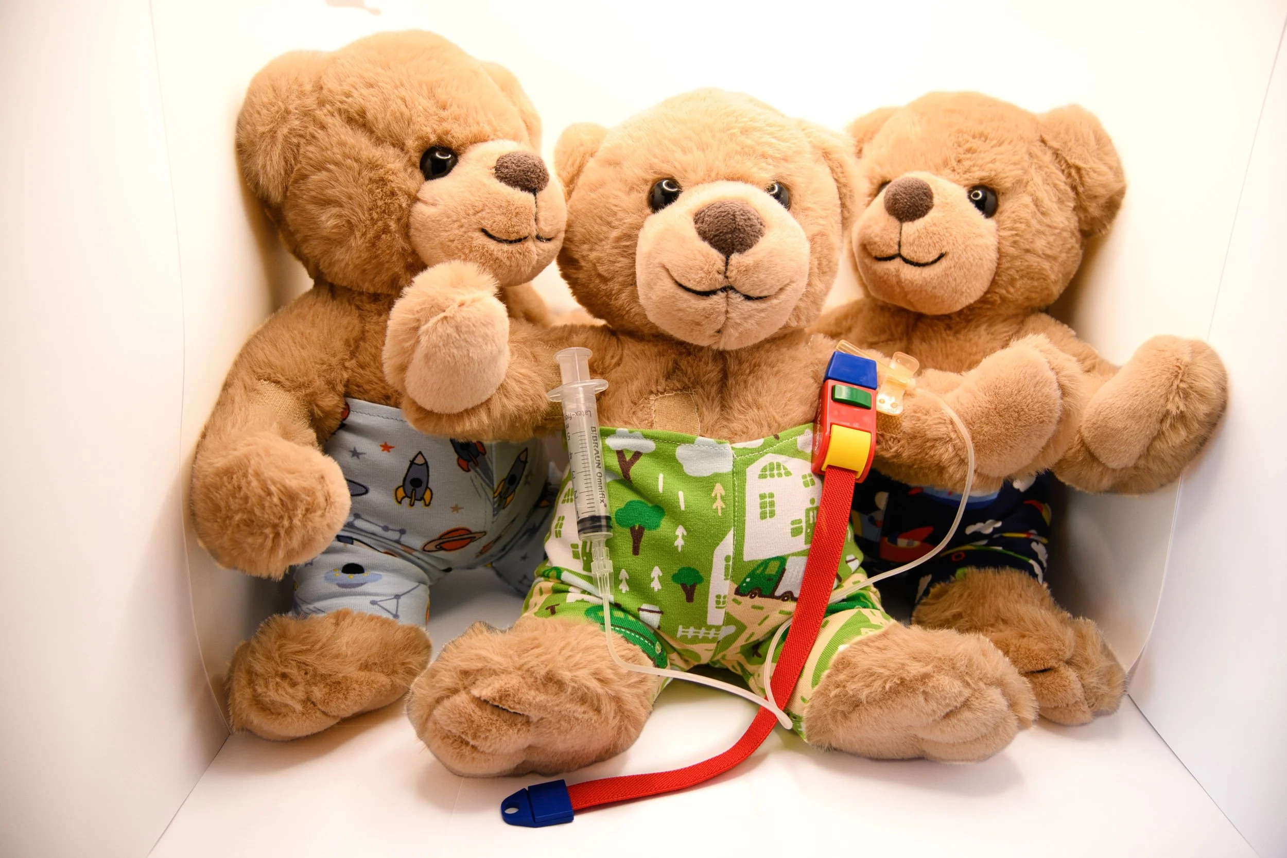 Three plush teddy bears dressed in colorful pajamas, each with medical instruments like a syringe and stethoscope, sitting together in a small white enclosure.