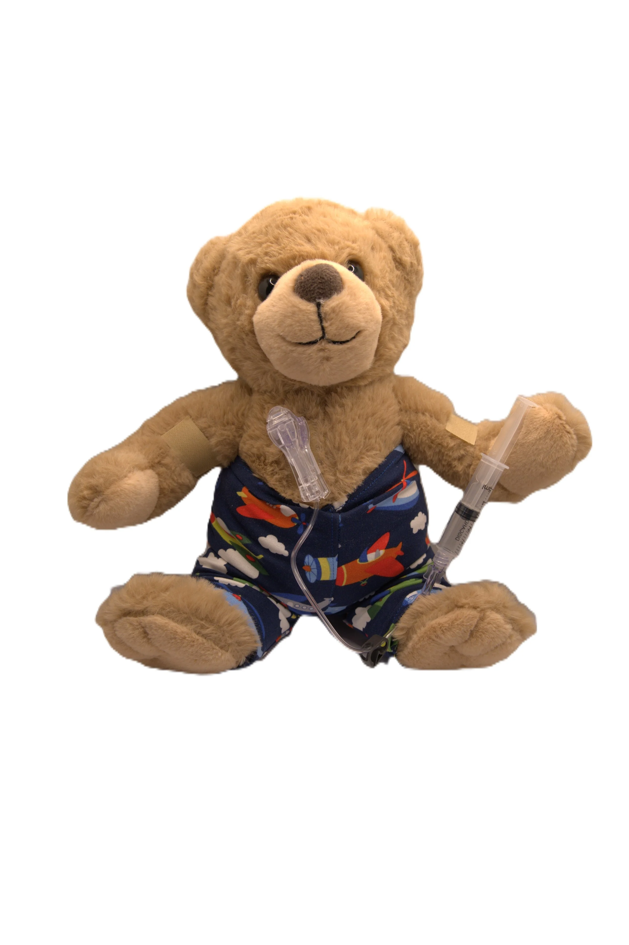 A plush teddy bear wearing a blue outfit with colorful rockets, holding a syringe and attached to a medical IV line.