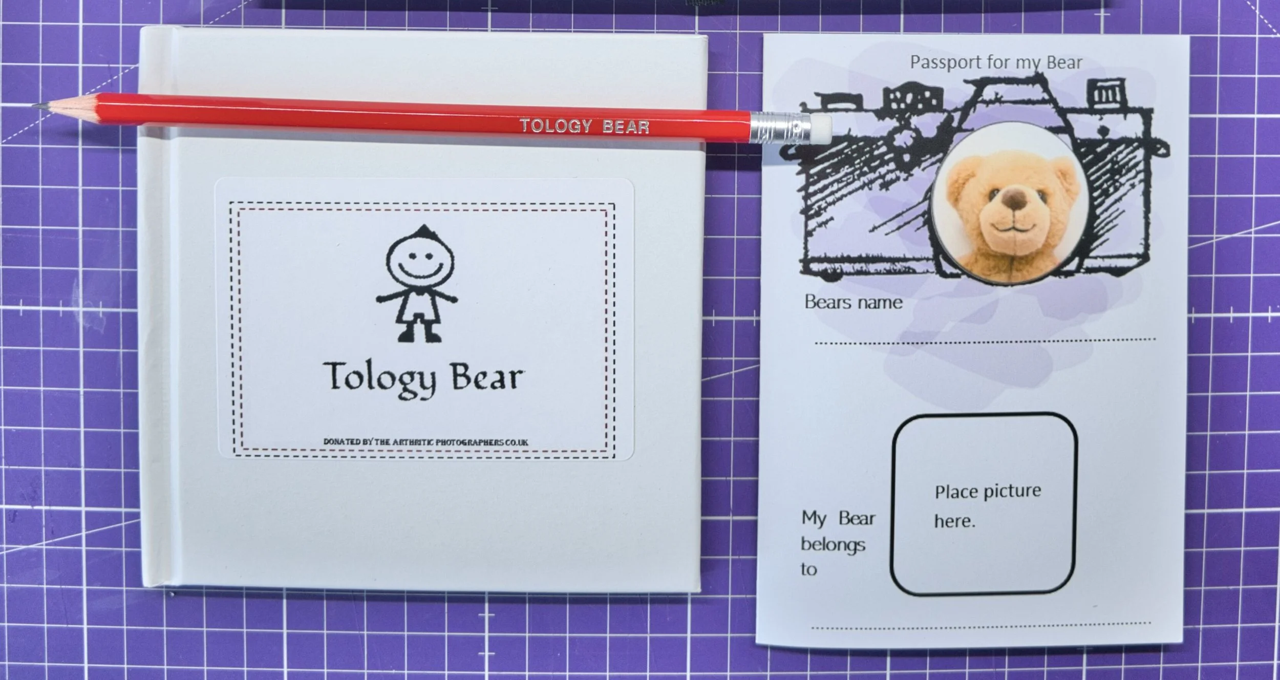 A purple cutting mat with a white booklet titled "Tology Bear" featuring a stick-figure drawing of a bear and the text "Donated by the Arthritic Photographers Co. UK." A red pencil labeled "Tology Bear" is placed on top of the booklet. To the right, there is a partially visible paper titled "Passport for my Bear" with a photo of a teddy bear and space for the bear's name and picture.
