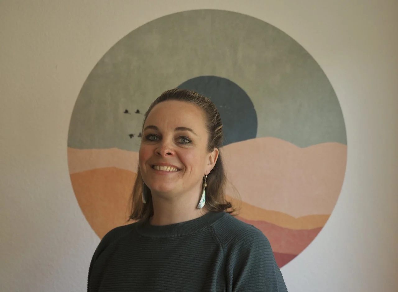 Frau steht vor einer Wand mit rundem, abstraktem Kunstwerk. Sie lächelt und trägt Ohrringe.