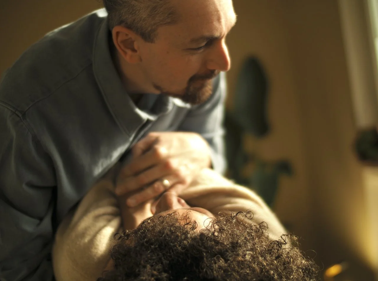 Ein Mann neigt sich über eine liegende Person, die ihn anschaut. Er hat einen Dutt  und einen Bart, während die liegende Person lockiges Haar hat. Der Hintergrund ist unscharf, mit Pflanzen angedeutet.