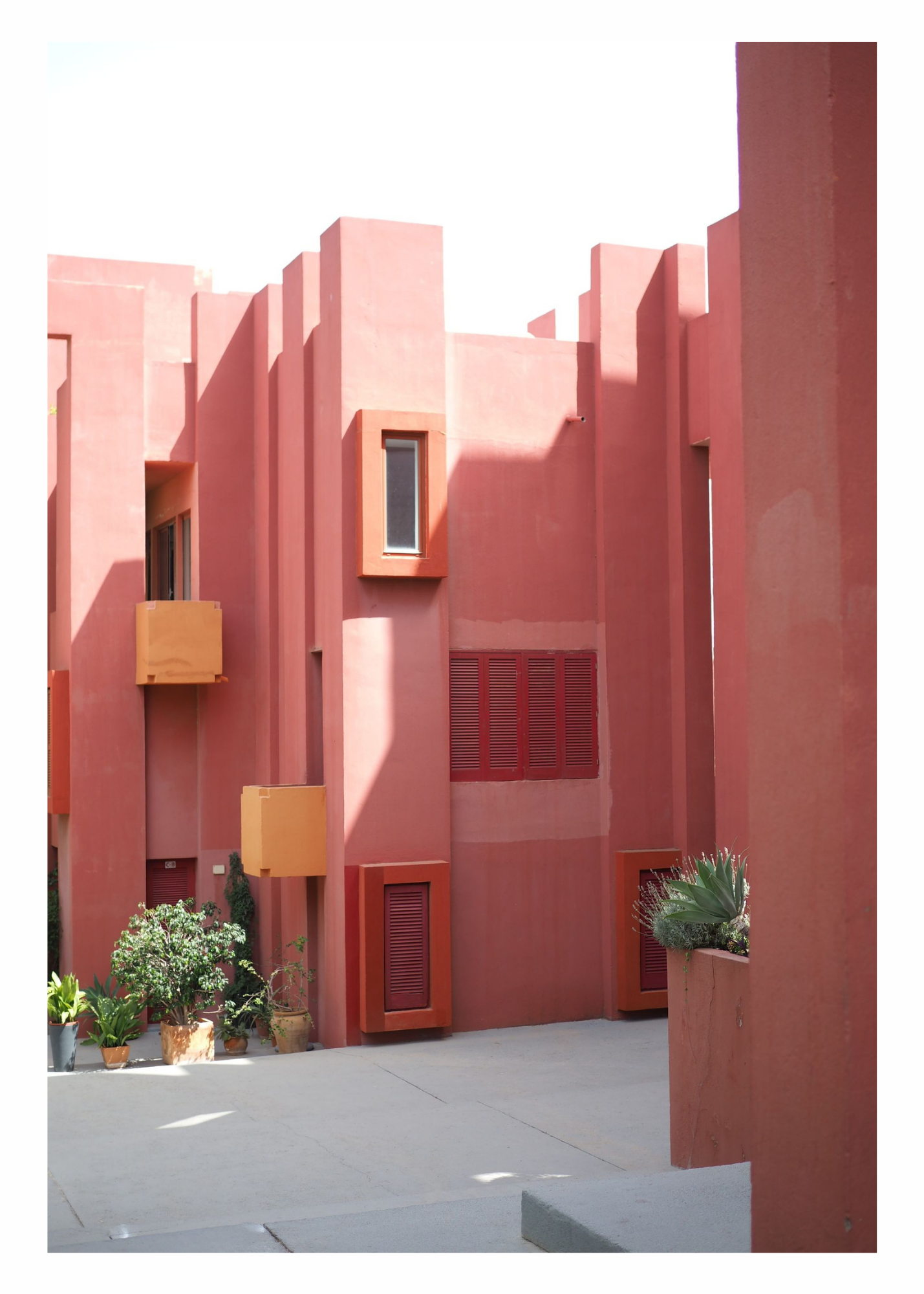 A pink stucco building with various small windows and rectangular architectural details. Potted plants are at the base of the building, and there's an open space in front of it with a concrete sidewalk.