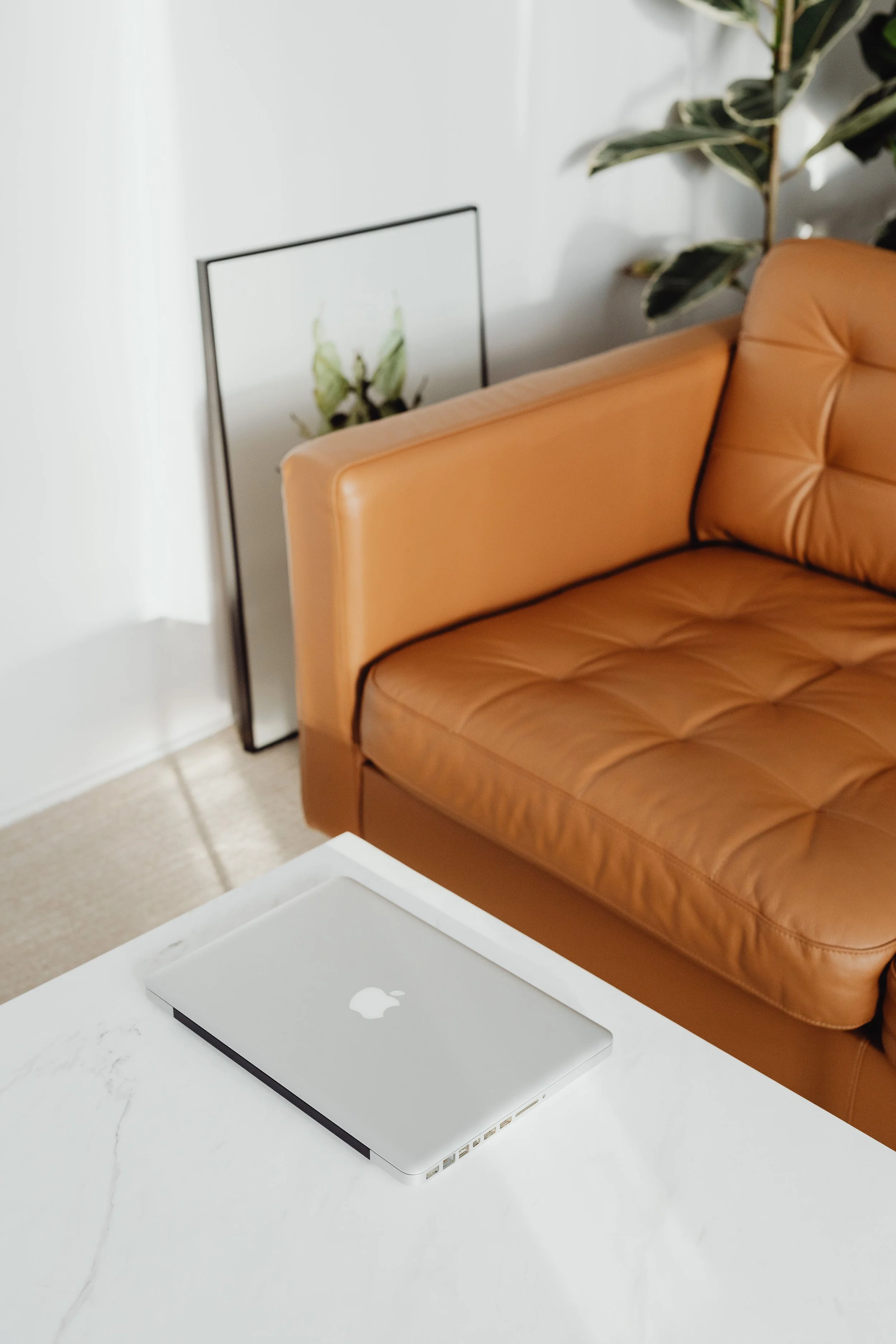 A closed silver MacBook laptop on a white marble table in a room with a tan leather sofa, a framed poster of a beetle, and a green plant with variegated leaves in the background.