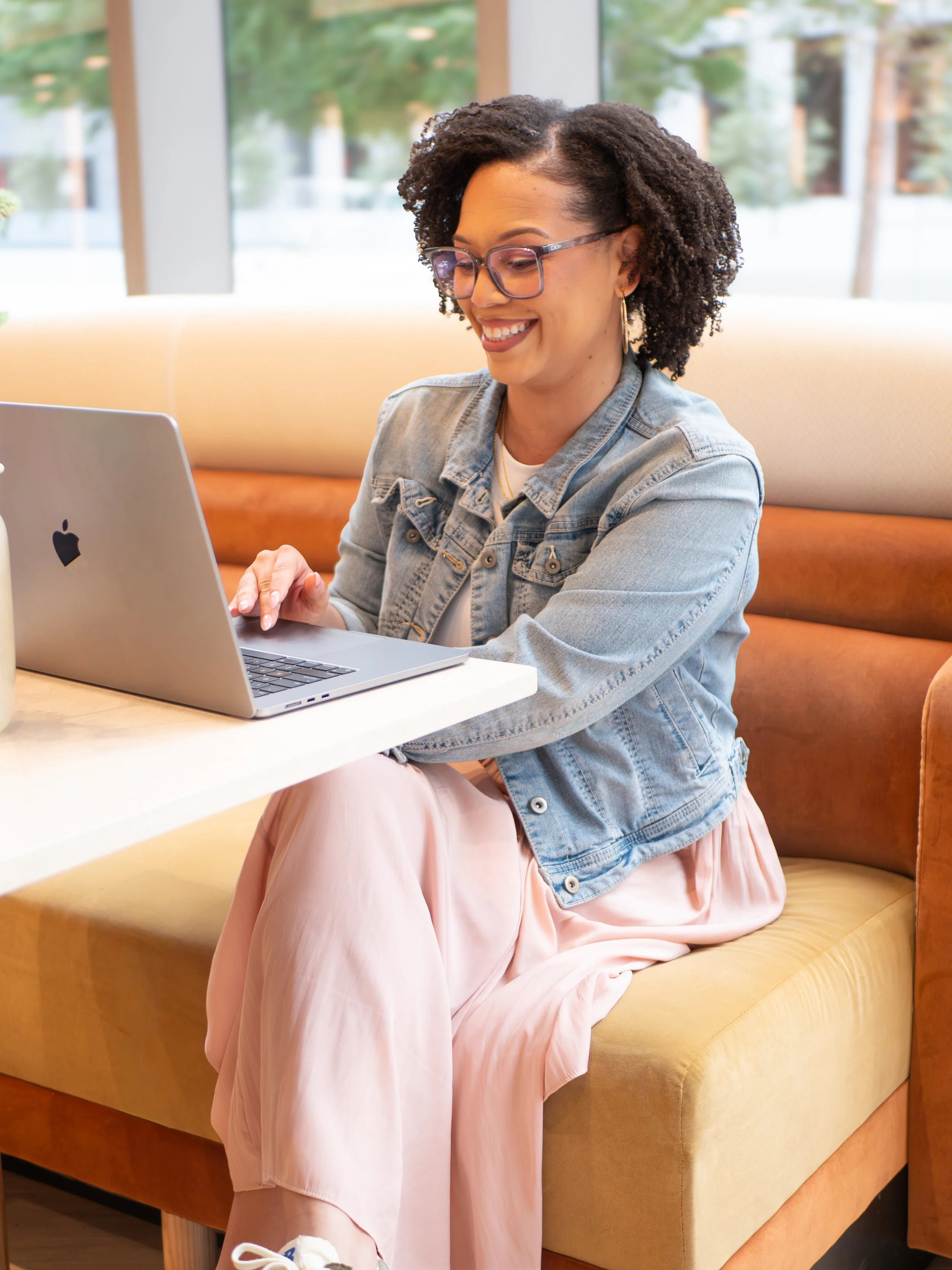 A young woman sitting on a yellow cushioned bench, working on a silver MacBook laptop, wearing glasses, a denim jacket, a white top, and a pink dress, in a bright room with large windows and green outdoor scenery.