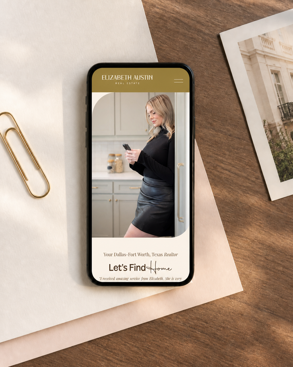 A smartphone displaying a real estate website with a woman using her phone in a kitchen. The website is for Elizabeth Austin, a Dallas-Fort Worth, Texas realtor, with the message "Let's Find Home." The phone is placed on a desk with papers, a gold paper clip, and a photograph of a building nearby.