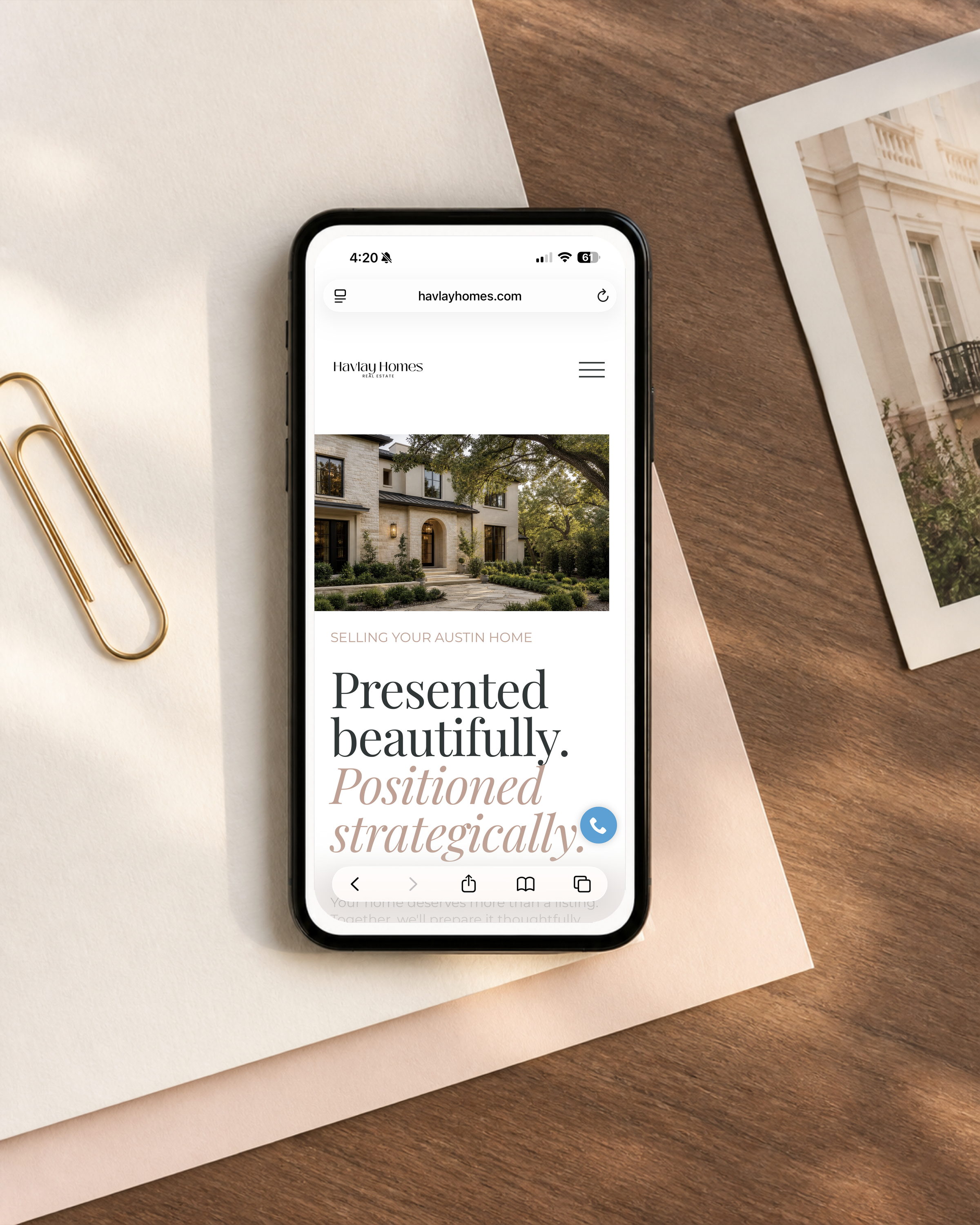 Smartphone displaying a real estate website with a large photo of a house, placed on pink and beige paper, with gold paper clips on the left.