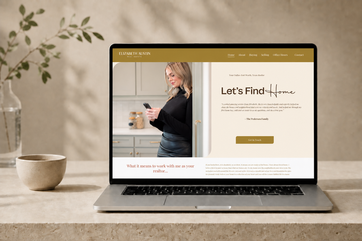 Open laptop displaying a real estate agent's website, with a woman in a black top and leather skirt looking at her phone, on a beige countertop with a plant in a glass vase and a ceramic bowl.