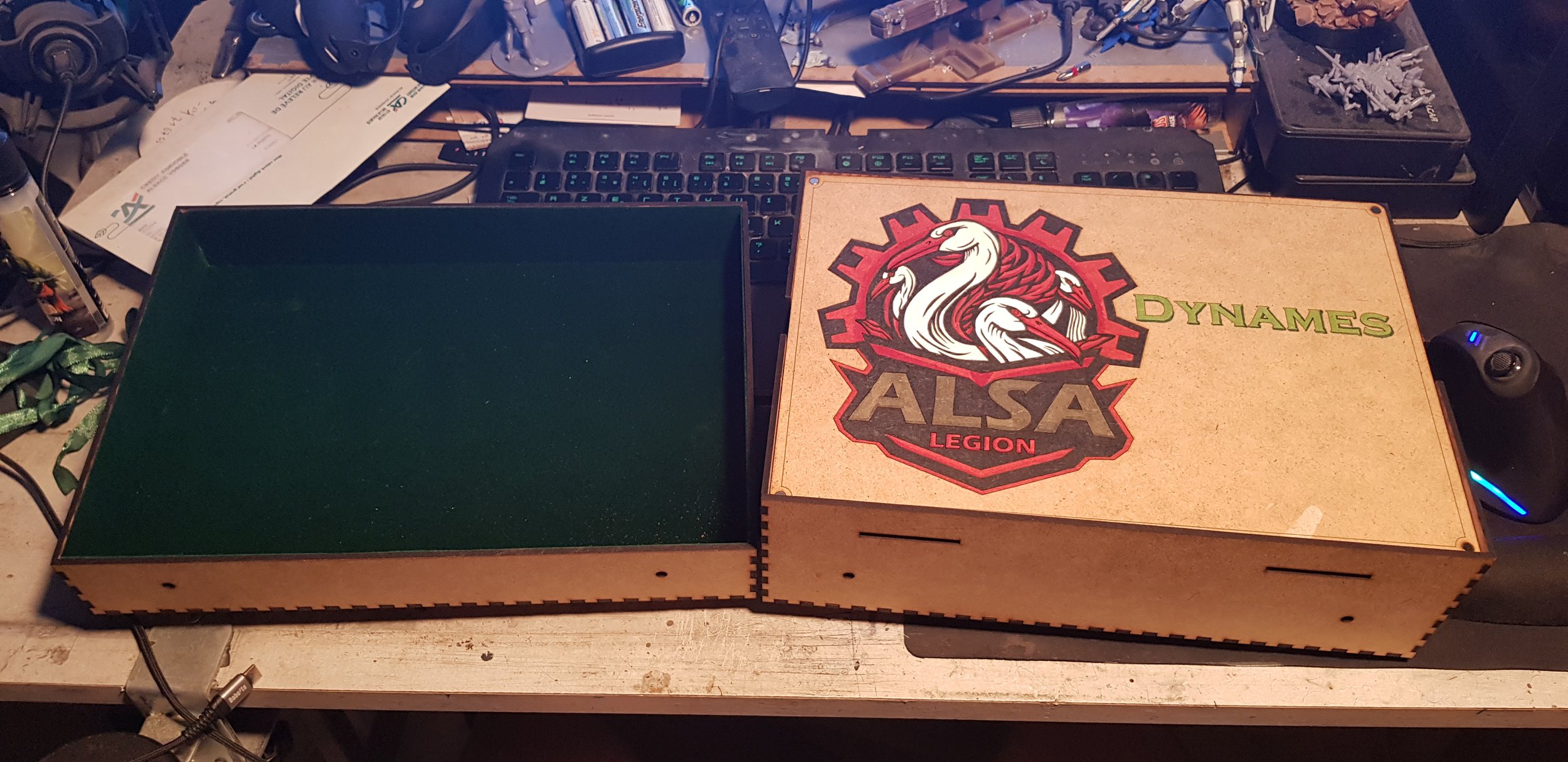 Deux boîtes en bois, une à gauche avec un intérieur vert, et une à droite décorée d'un logo avec un cygne et le texte 'ALSA LEGION' et 'DYNAMES', sur un bureau avec un clavier, un casque, une souris et divers objets.
