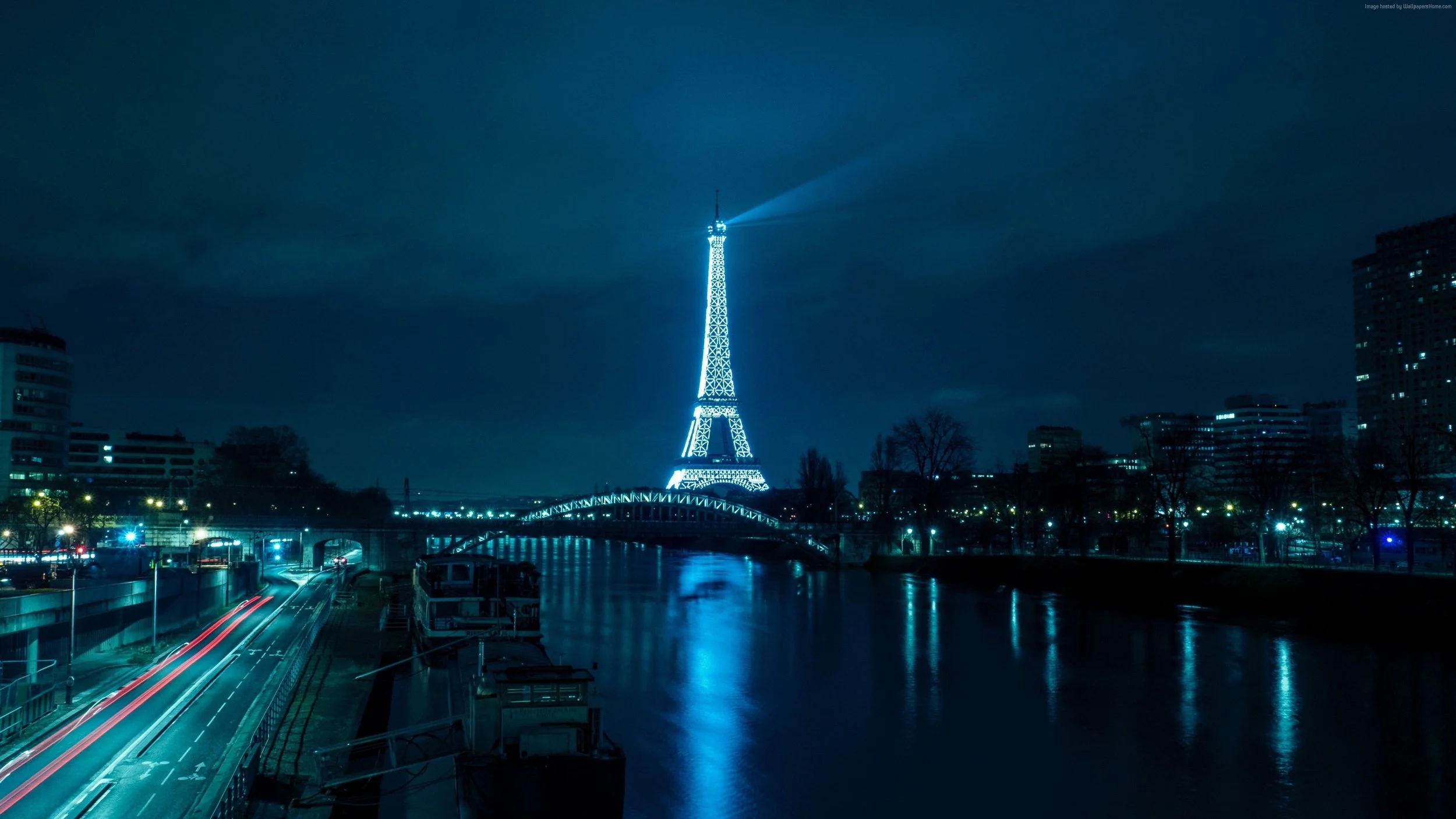 La Tour Eiffel illuminée la nuit avec reflection dans la Seine et un pont, en arrière-plan des bâtiments modernes et un ciel nuageux.