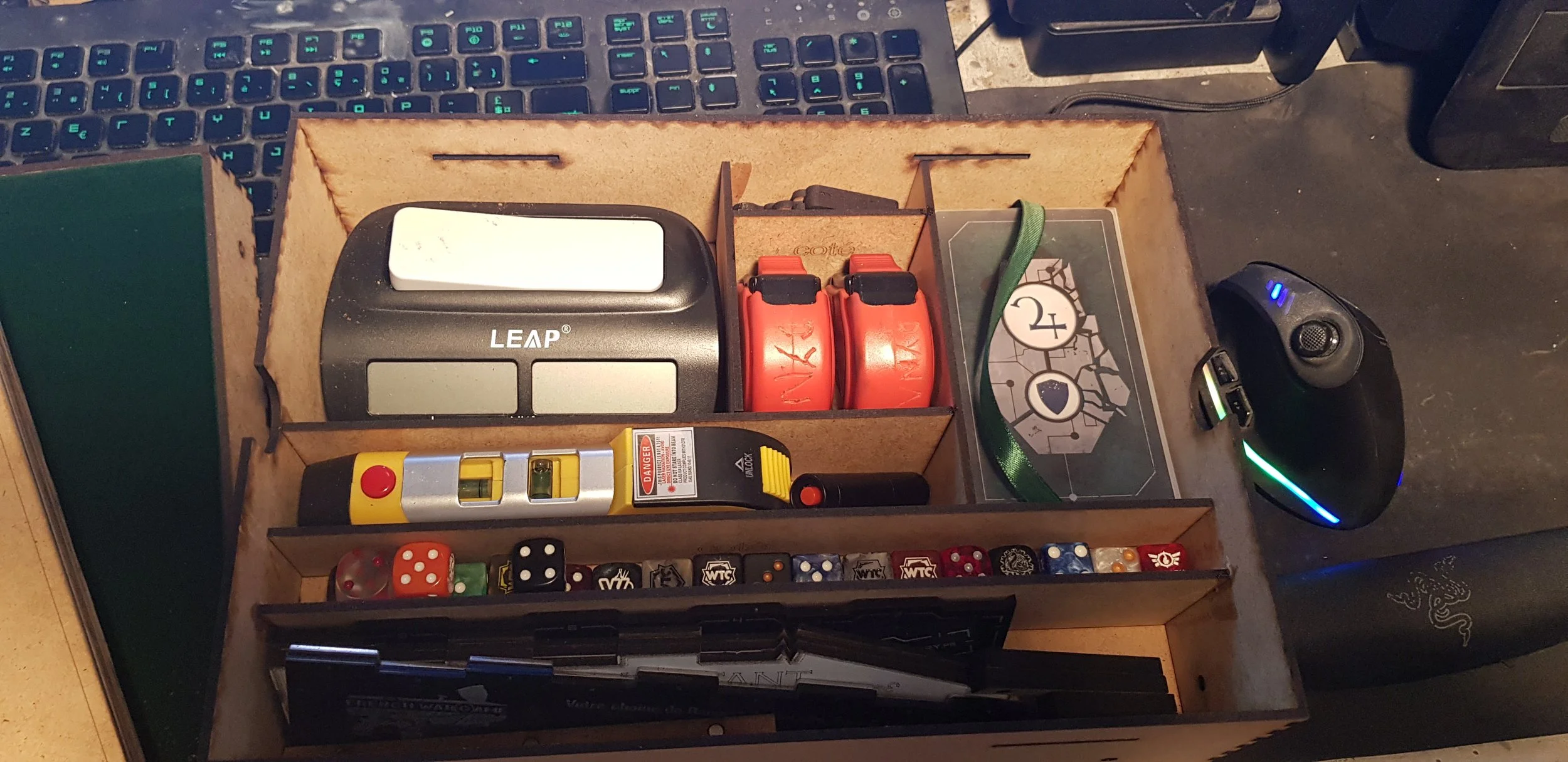 Une boîte à outils en bois contenant divers objets : un outil de mesure laser, des rubans à mesurer rouge, une boîte de rangement, des dés, un clavier d'ordinateur en arrière-plan, une souris de jeu avec éclairages LED, et un écran vert