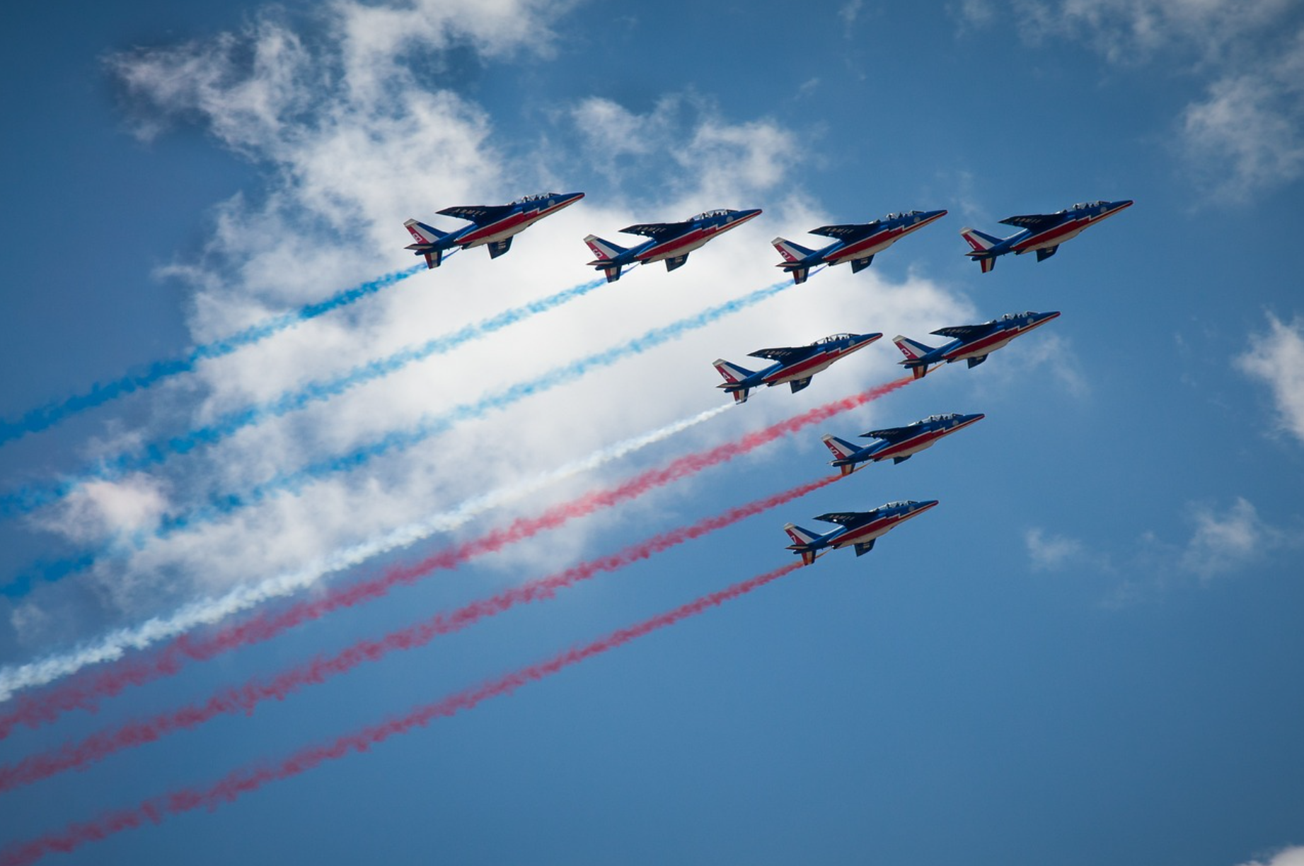 Huit avions de chasse colorés en formation, laissant des traînées de fumée tricolores françaises dans le ciel bleu avec quelques nuages.
