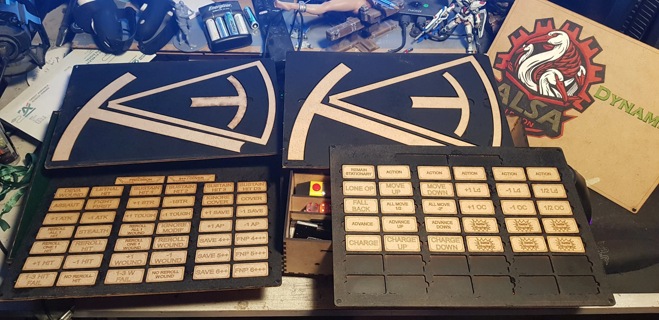 Table de jeu de rôles avec des cartes et des figurines, entouré de diverses piles et accessoires sur une surface de bois, avec un logo de dragon blanc et rouge sur une boîte en carton, et une plaque avec de nombreux boutons