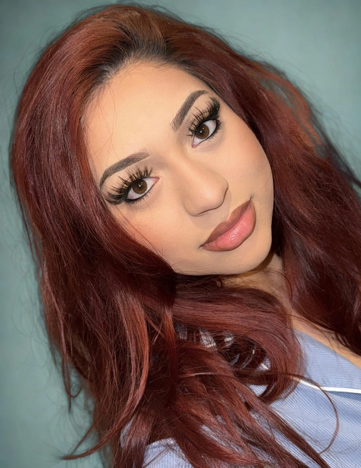 A woman with long, wavy red hair, makeup with defined eyebrows, long eyelashes, and wearing a blue top, taking a selfie indoors.