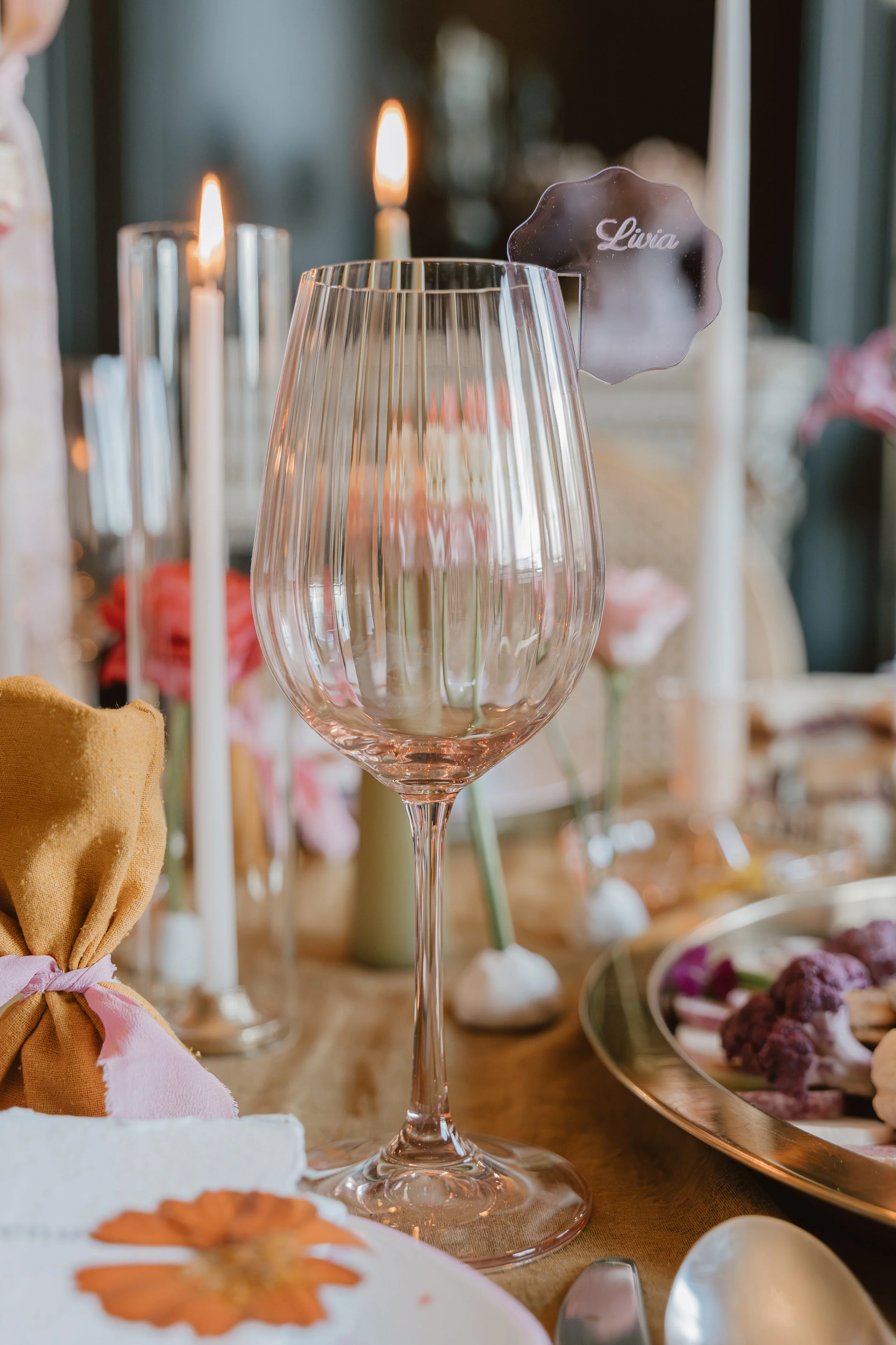 Verre à vin en verre transparent, décoré d'une étiquette portant le nom 'Livia', placé sur une table de fête avec des bougies allumées et des fleurs roses en arrière-plan.