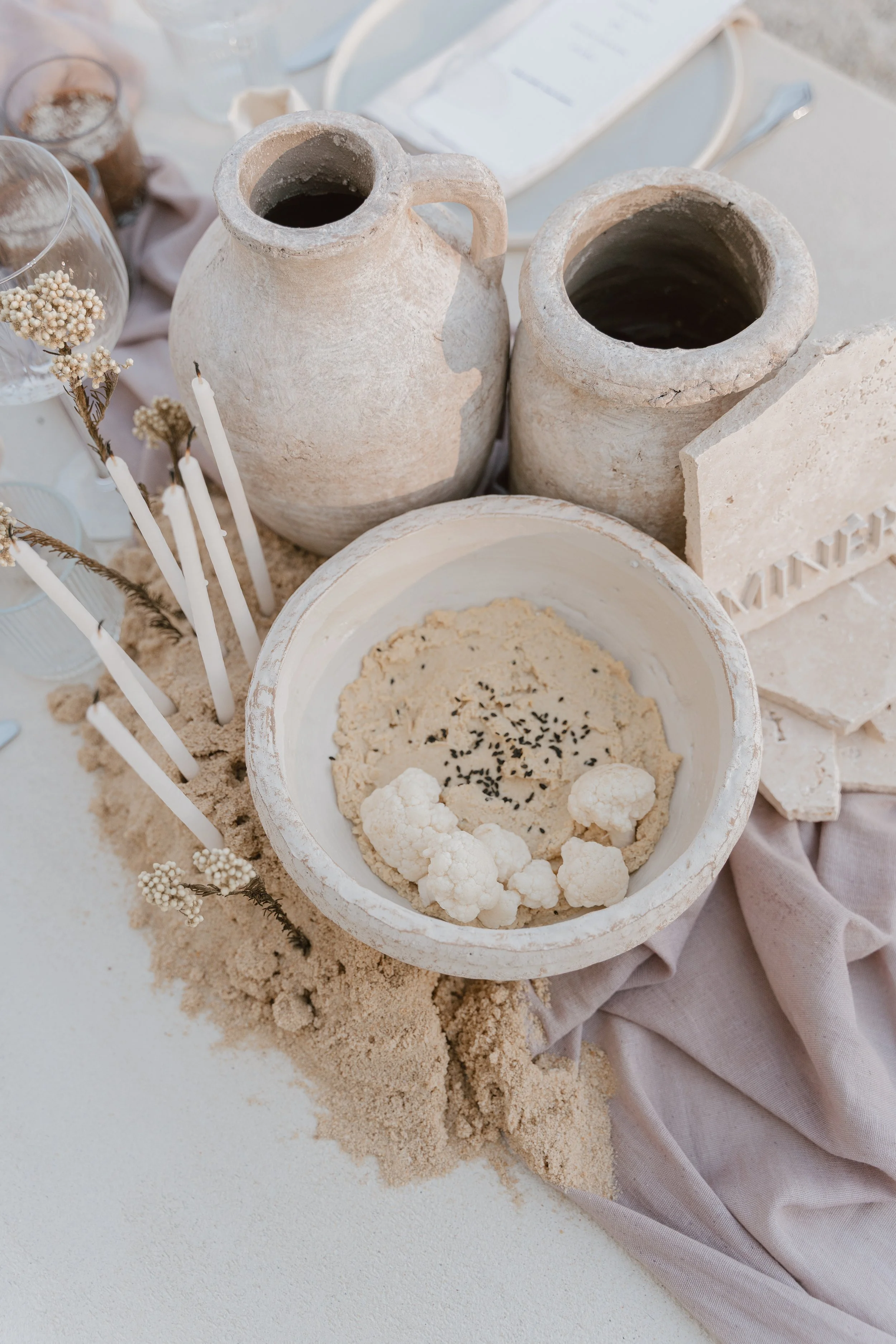 Décor de table avec des jars en terre cuite, une assiette de hummus avec des morceaux de chou-fleur, bougies blanches, fleurs séchées, et plaques de pierre sur une nappe beige.