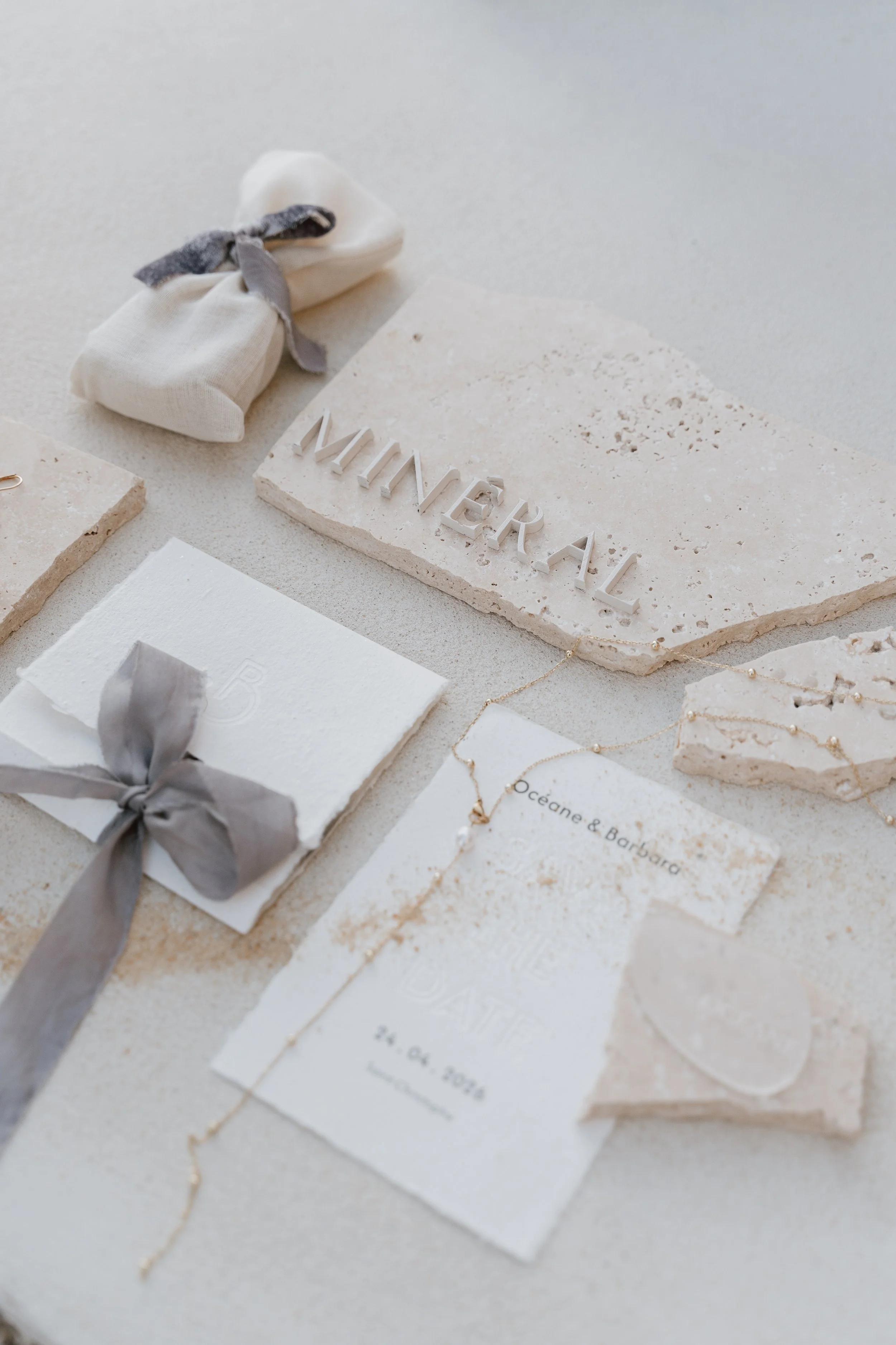 Invitation de mariage avec des éléments en pierre, une carte de vœux blanche avec un ruban gris, un collier délicat, et une pierre en forme de cœur, tous disposés sur une surface claire.
