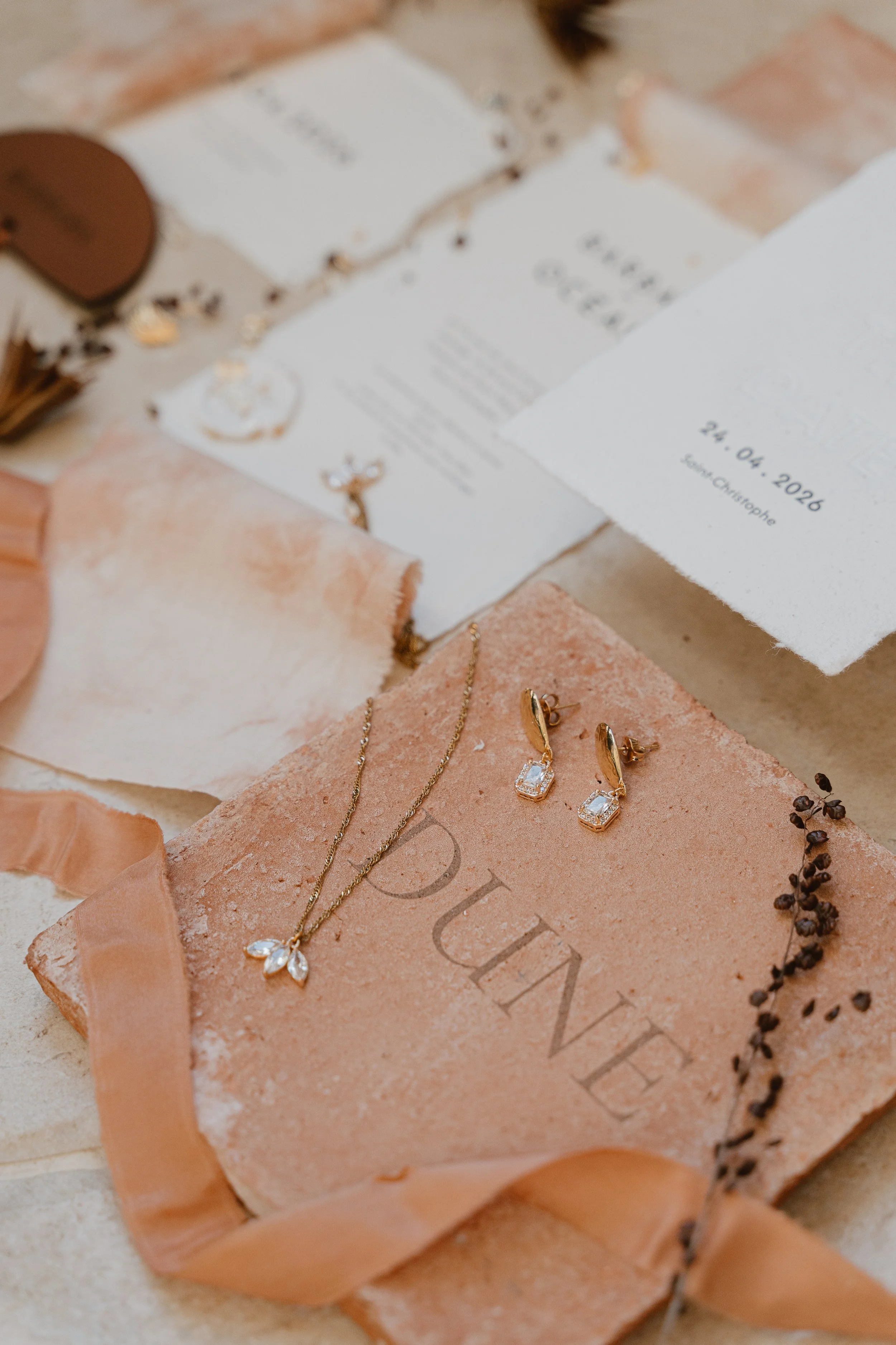 Collection de bijoux et d'invitations de mariage sur une surface en pierre, comprenant un collier, des boucles d'oreilles, une carte avec la date 24 avril 2020 et un lieu, et un livre avec le mot 'PLUME' en relief.