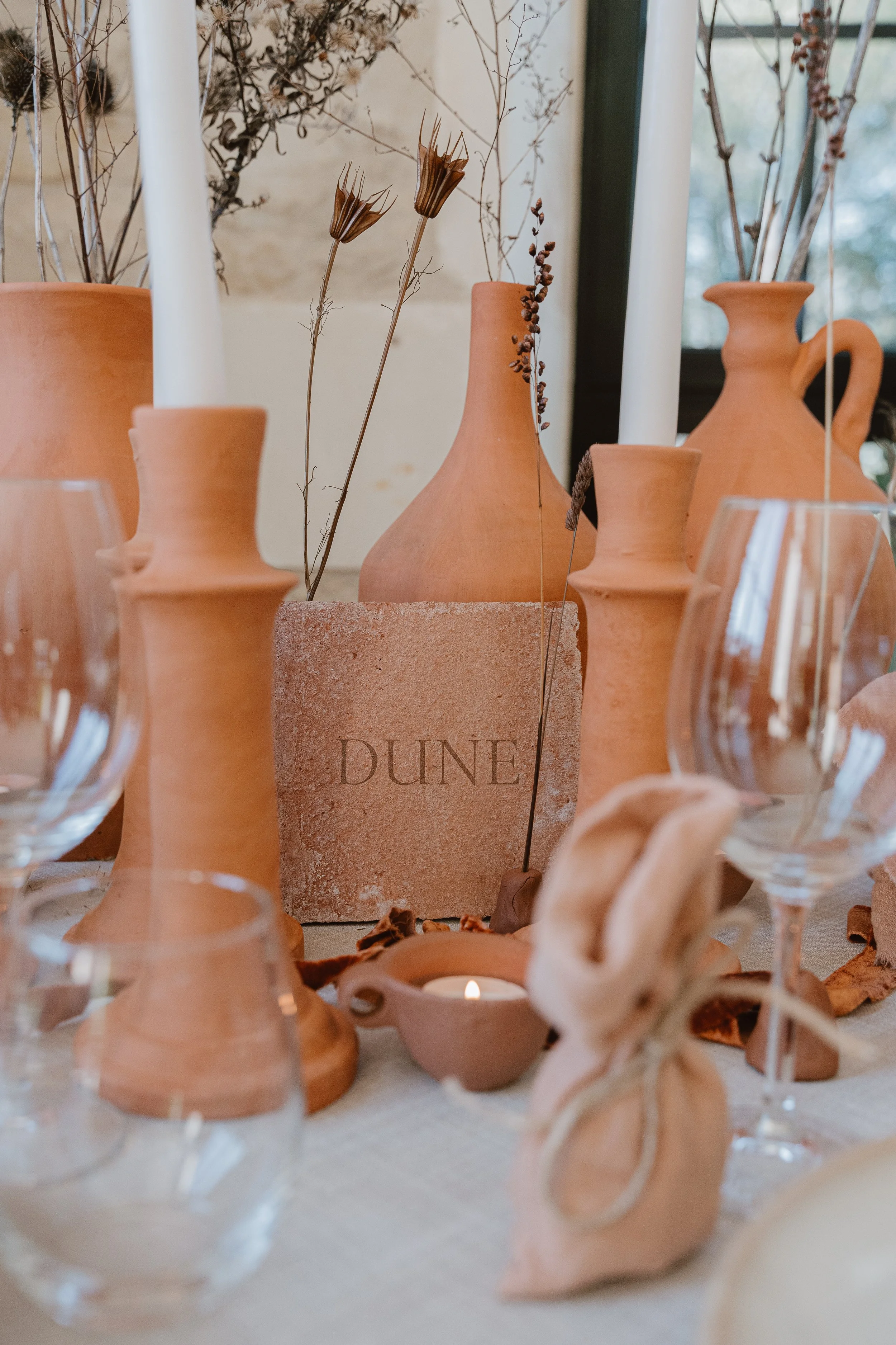 Vase en terre cuite, bougeoir et verres à vin, décor minimaliste avec branchages secs et une pierre gravée 'DUNE' sur une table en tissu beige.
