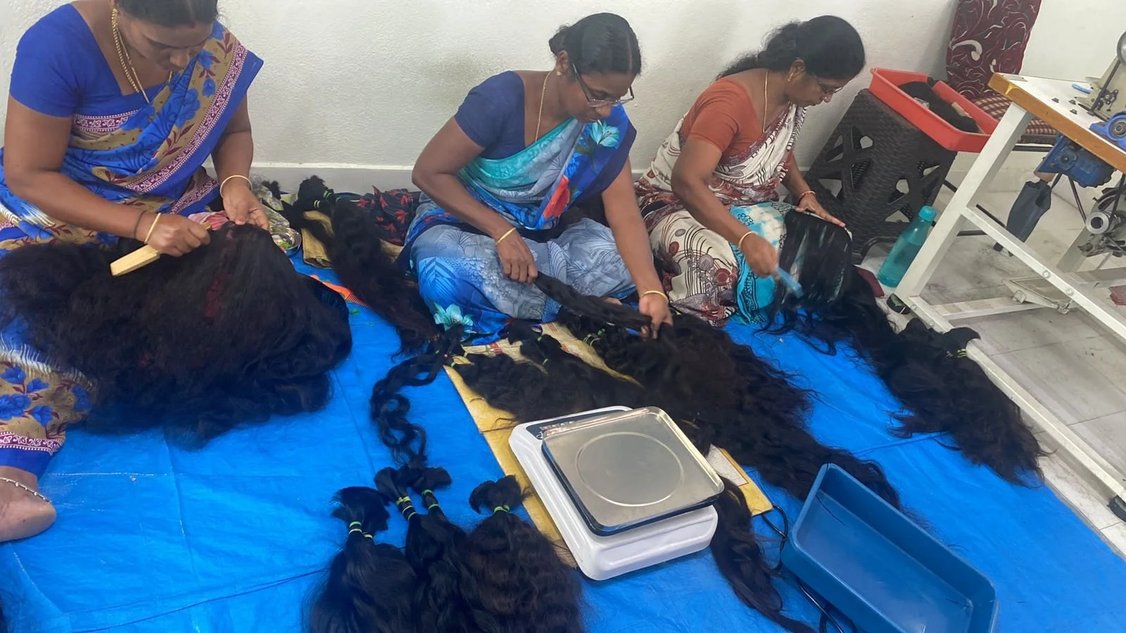 Three women seated on the floor working on weaving long black hair extensions with colorful bands, surrounded by hair bundles and tools.