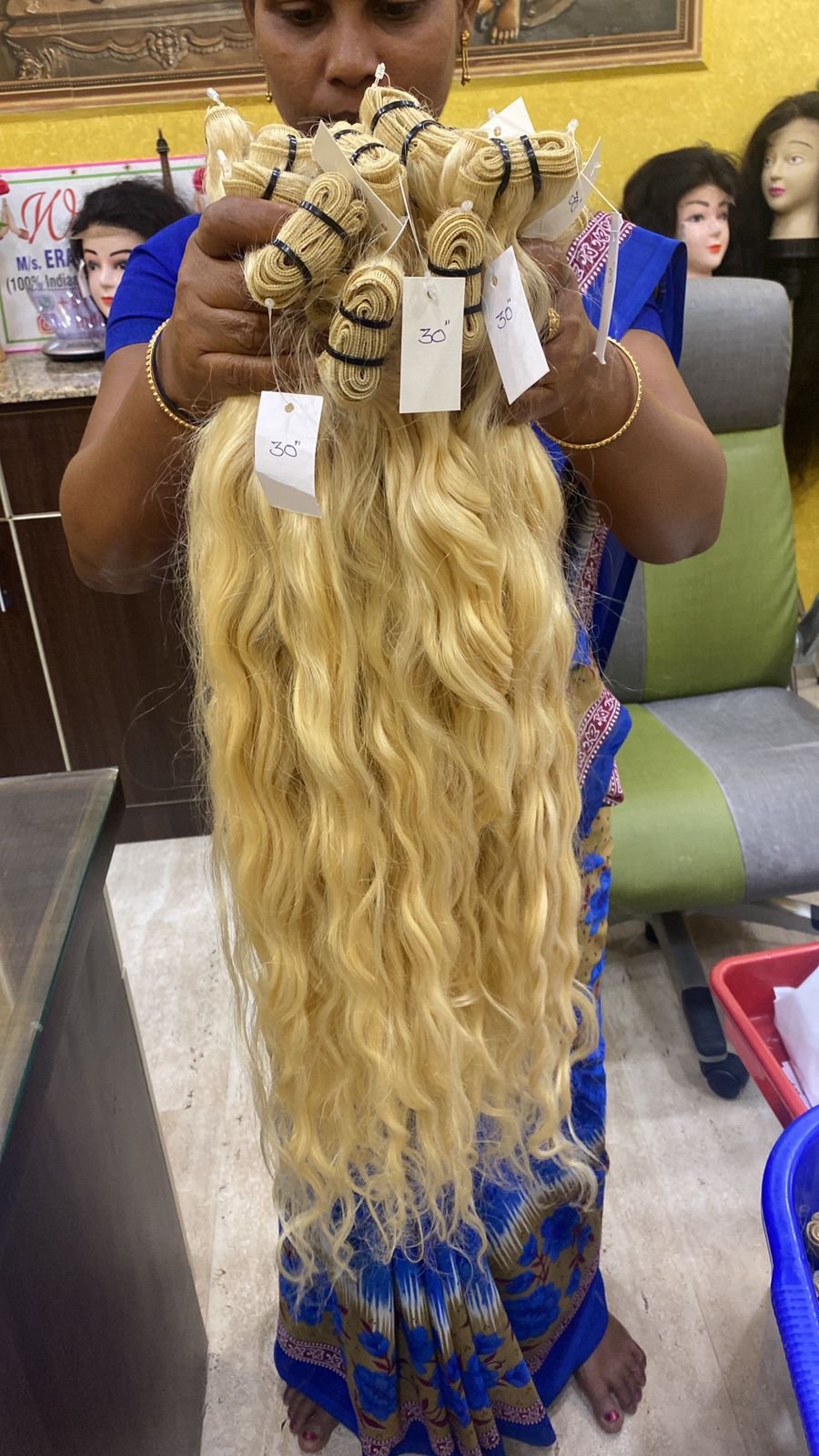 A woman holding a bundle of long, wavy, blonde hair extensions with tags indicating lengths of 30 and 50 inches in a wig shop filled with mannequin heads.