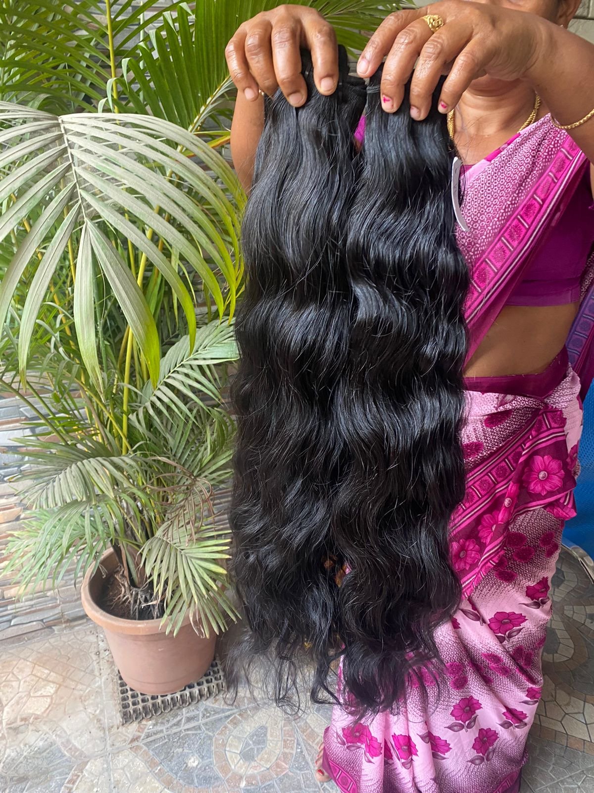 A woman in a pink sari holding a long, wavy black hair extension or wig outdoors with green plants in the background.