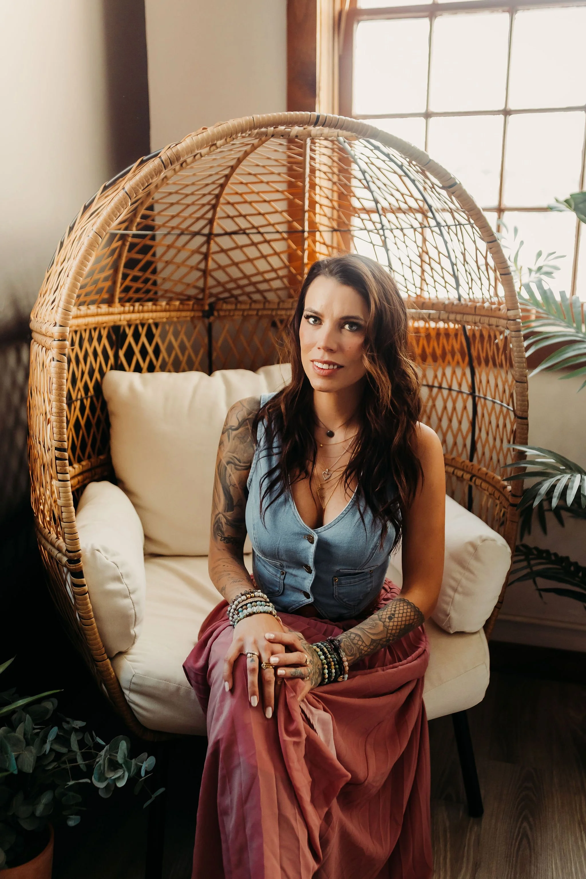 A woman with long wavy brown hair and tattoos on her arms is sitting on a cream-colored cushioned chair with a wicker canopy around her. She is wearing a sleeveless denim top and a long skirt, with multiple bracelets and rings. She is looking at the camera with a slight smile. There is a large window with wooden trim behind her, and some green plants are visible around.