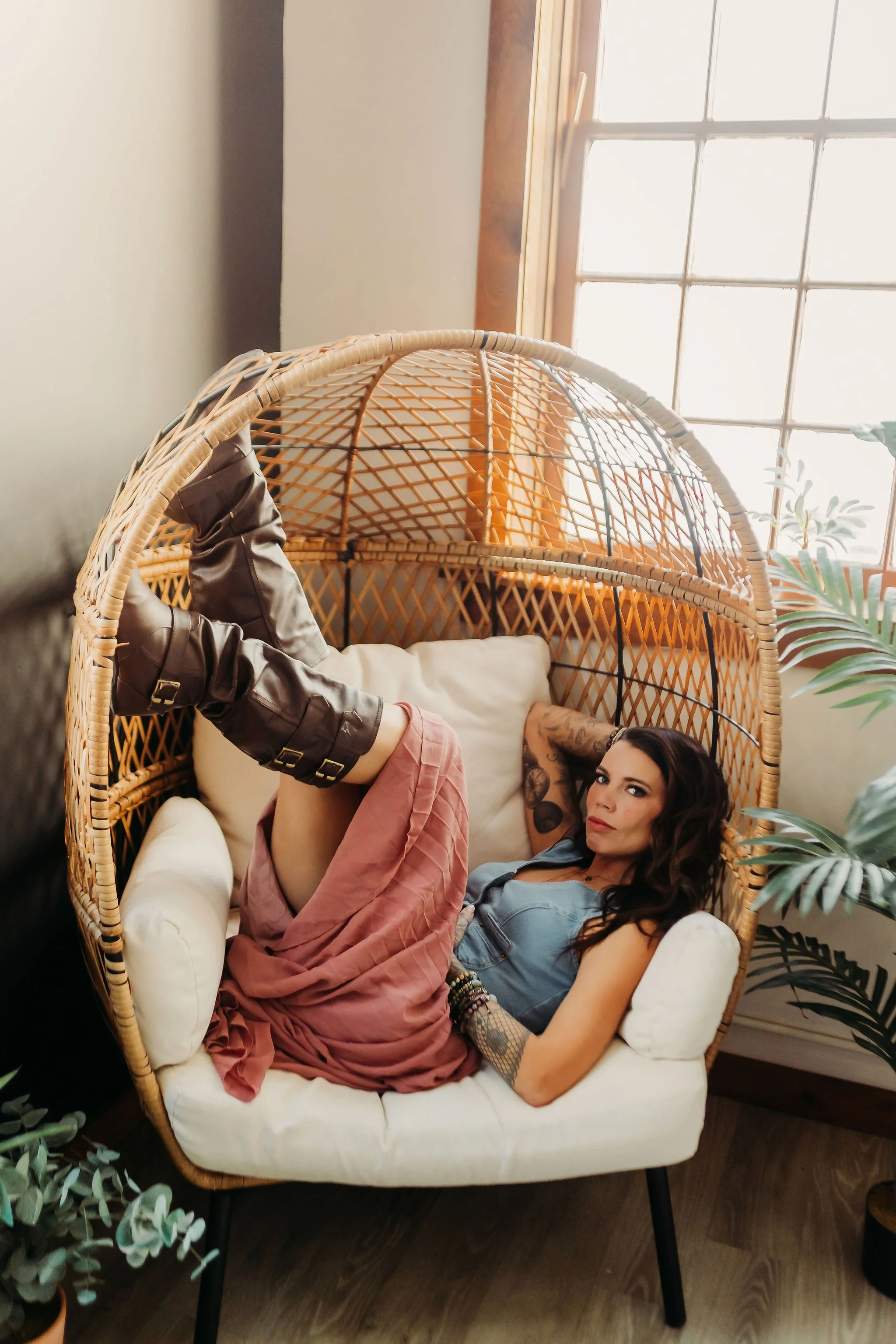 A woman with dark wavy hair, tattoos on her arm, and wearing a denim top and pink skirt, is lying inside a wicker hanging chair by a window with sunlight streaming in.