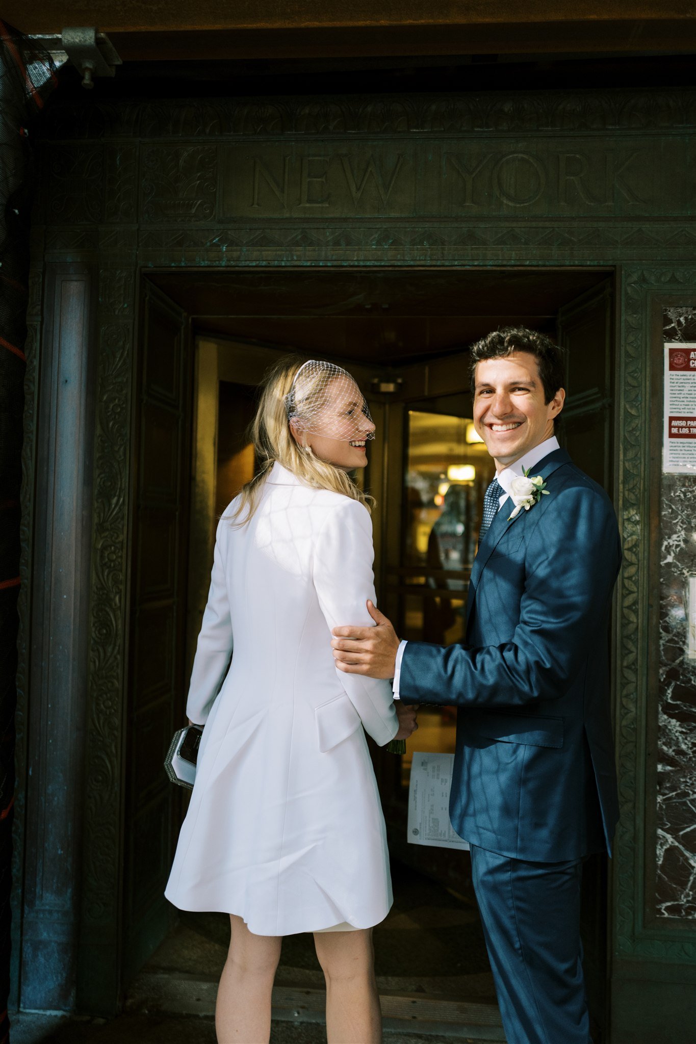 The couple smiles at a vintage doorway marked 'NEW YORK' before stepping inside. nyc courthouse wedding michelle rago destinations.