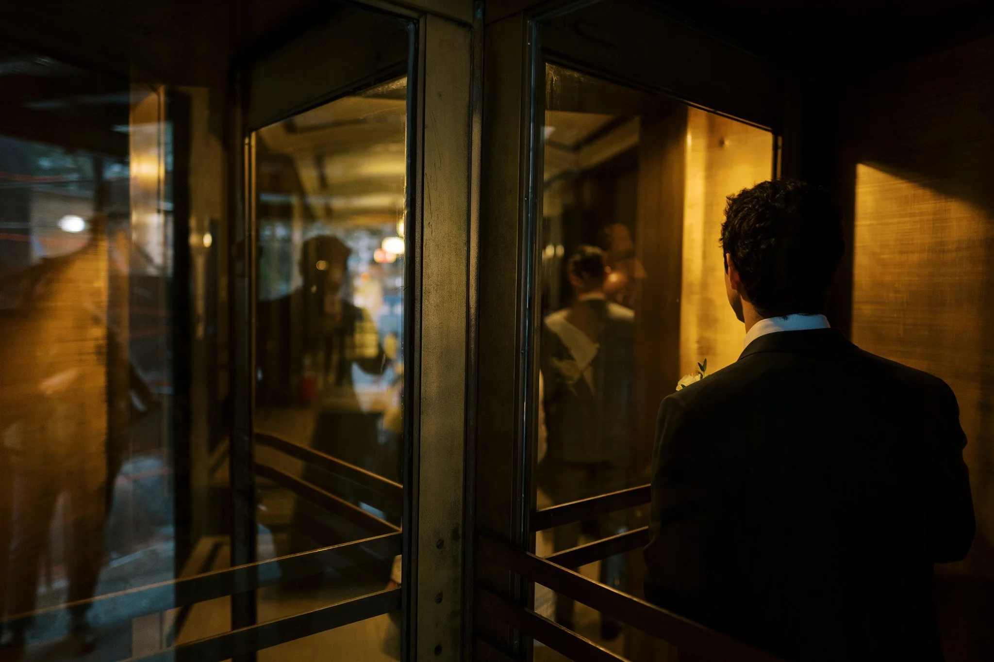 Groom steps through a revolving lobby door with warm light inside. the plaza hotel nyc luxury wedding photographer.