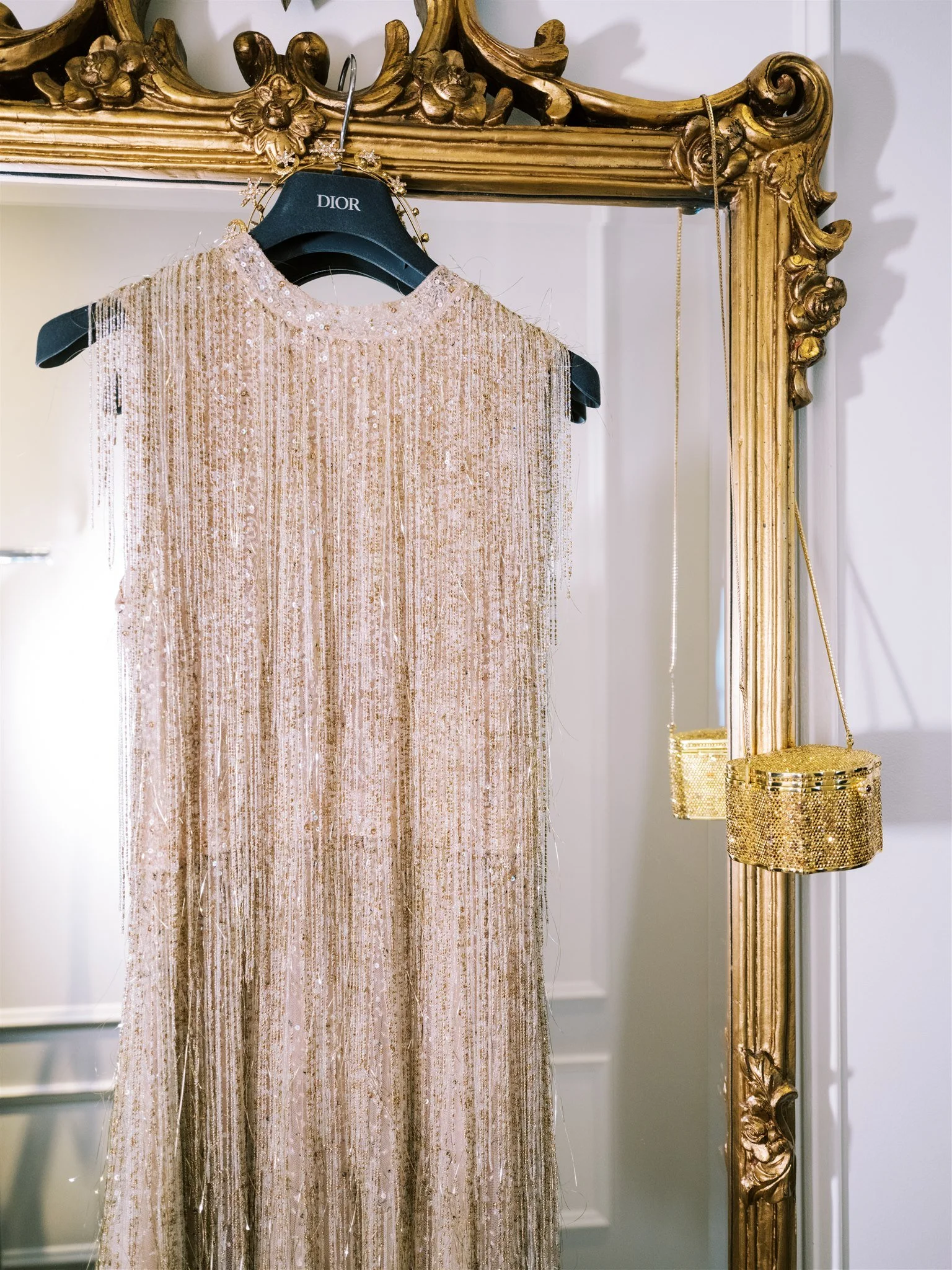 Gold‑fringe Dior dress and beaded bag hang from an ornate mirror. new york city luxury wedding photographer.