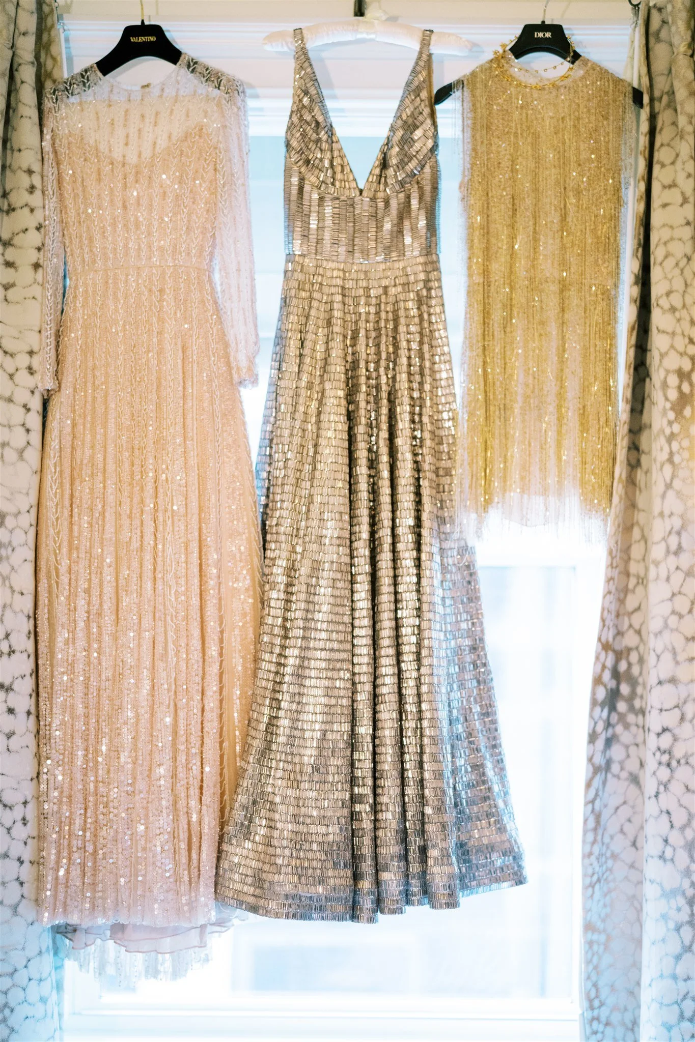Three shimmering evening gowns hang in a window between drapes. michelle rago destinations wedding planner nyc.