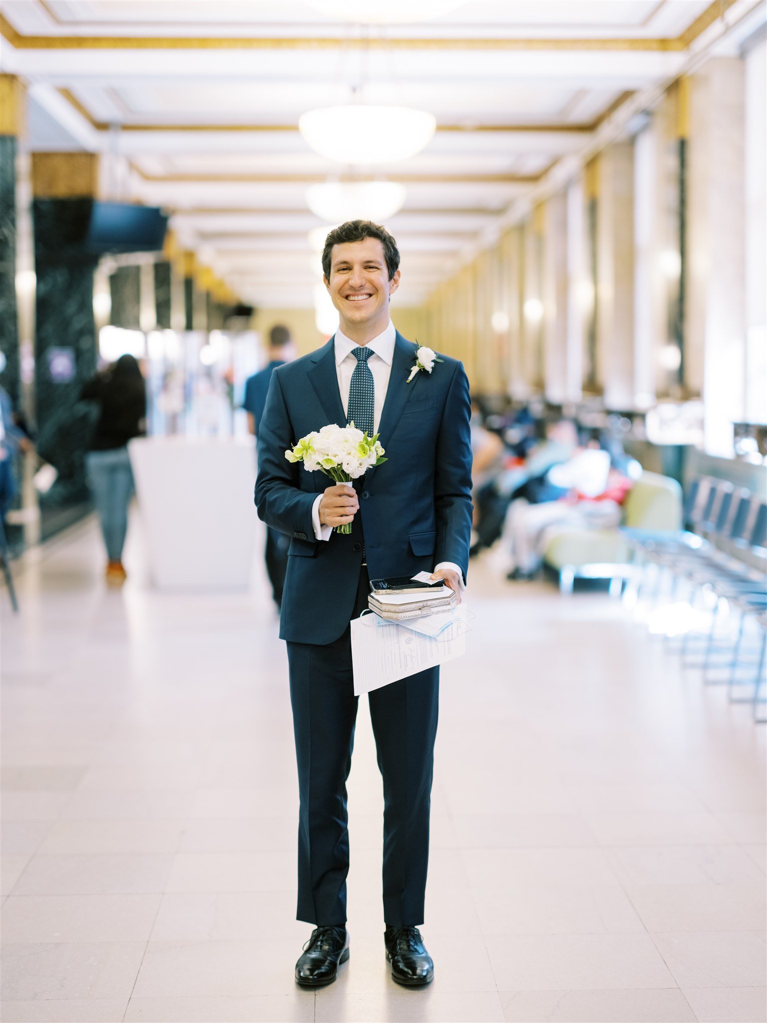 Groom in a navy suit holds a small bouquet while waiting in a bright courthouse hall. new york city courthouse elopement photography.