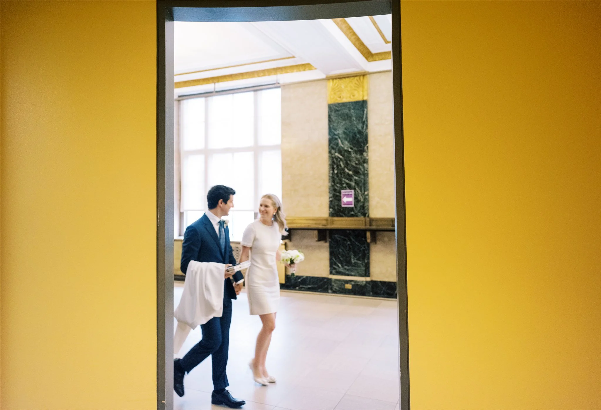 The couple walks past a yellow doorway in the courthouse captured mid‑stride. the plaza hotel new york destination wedding.