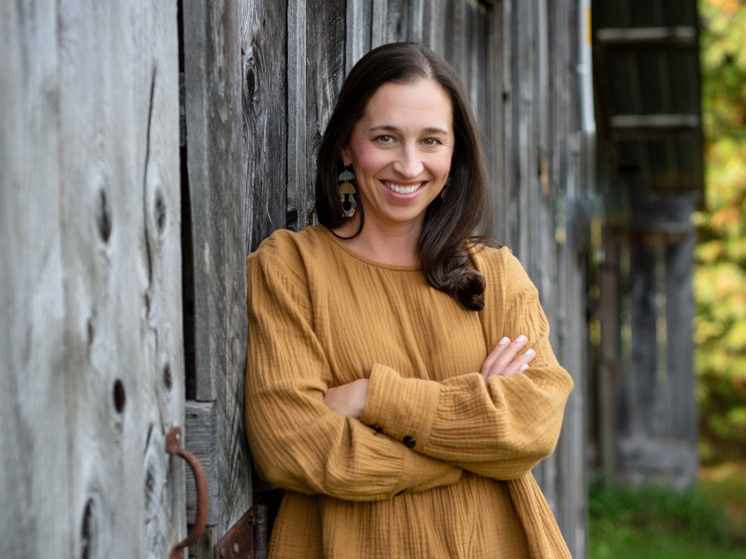 A woman with long dark hair, smiling, wearing earrings and a mustard-colored sweater, leaning against a wooden wall outdoors.