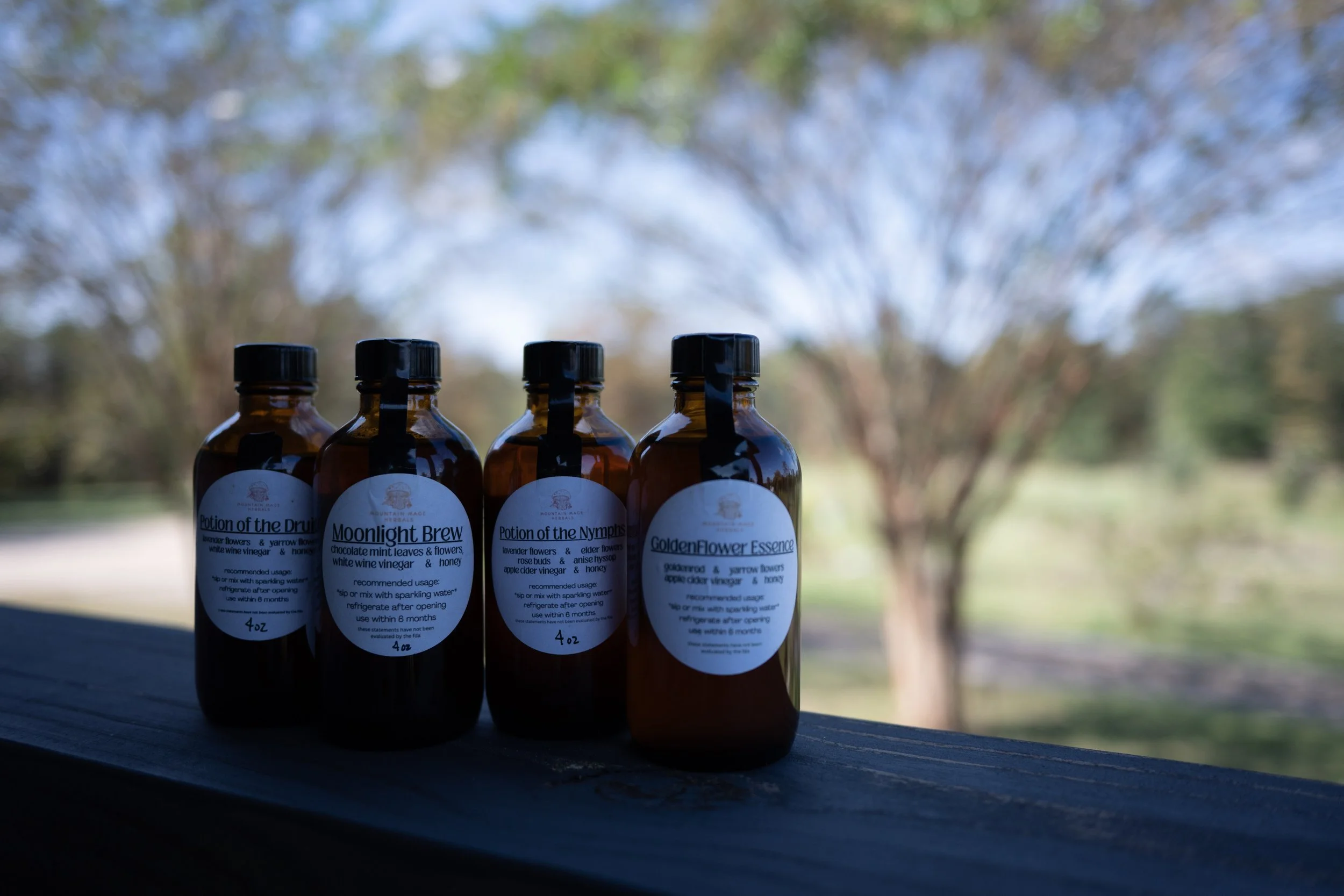 Four amber glass bottles with white labels and black caps sit outdoors on a flat surface, with a blurred background of trees and sky.