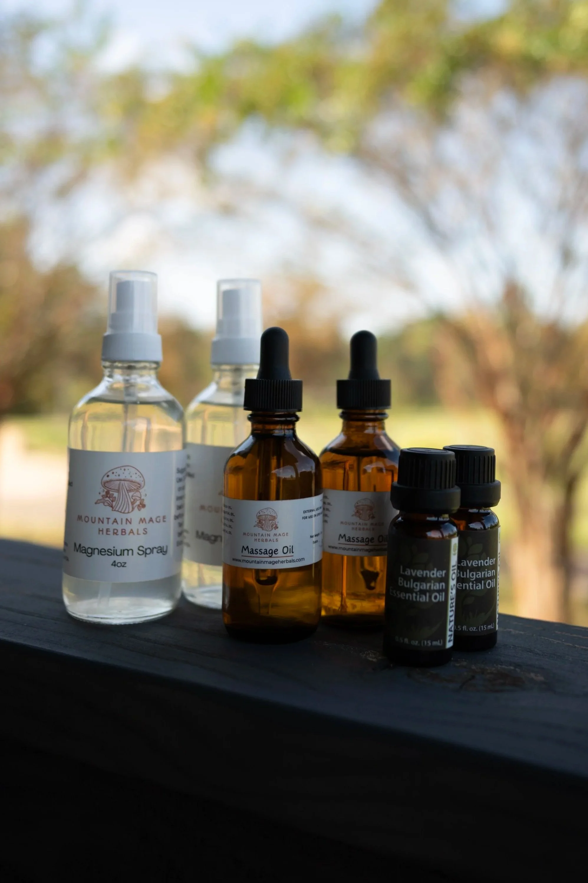 Multiple bottles of herbal remedies and essential oils, including magnesium spray, massage oil, and lavender Bulgarian essential oil, arranged on a wooden surface outdoors with a blurred background of trees and sky.