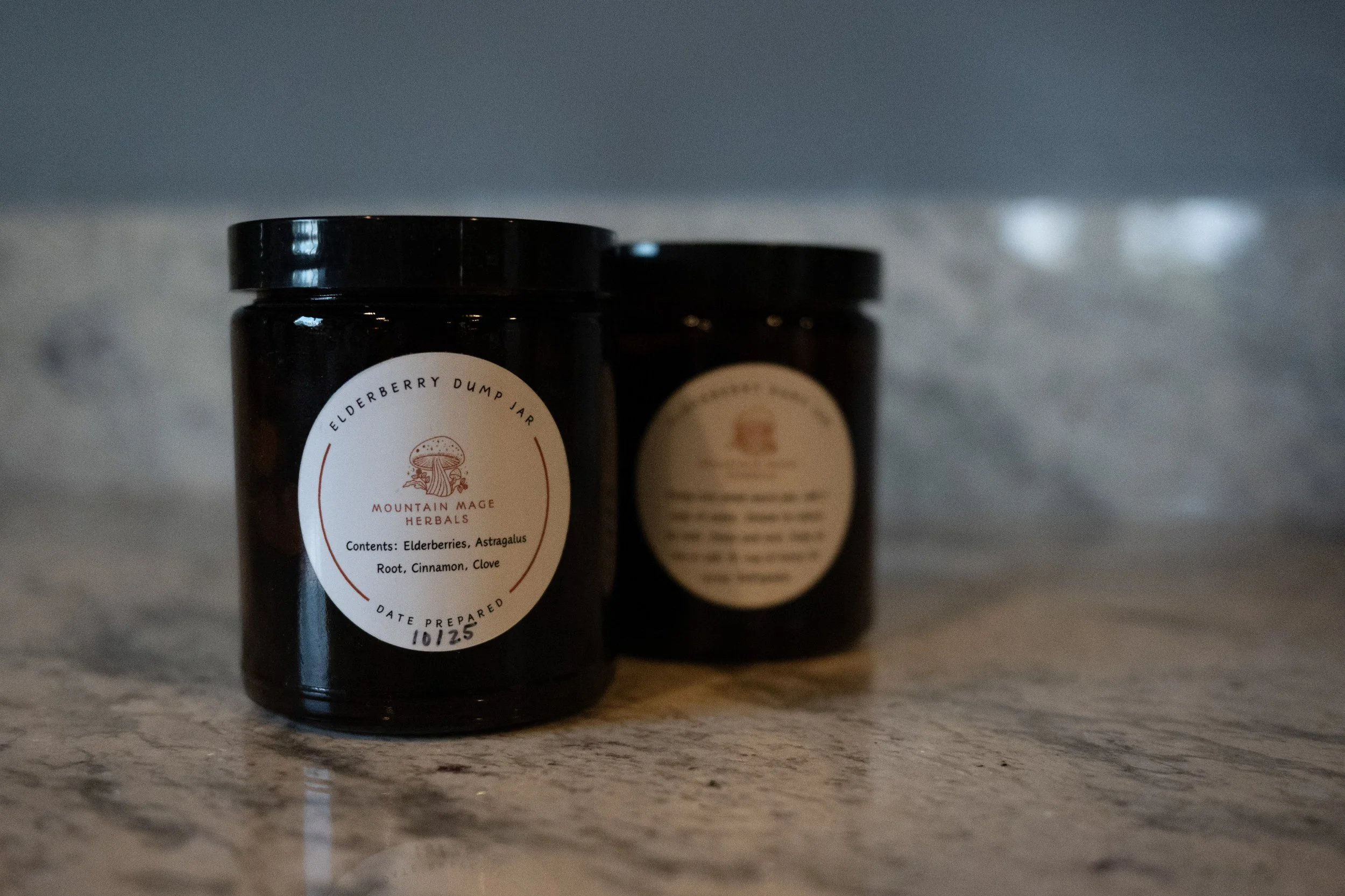 Close-up of two black jars, one in focus, labeled Elderberry Dump Jar from Mountain Mage Herbals, with ingredients like elderberries, astragalus root, cinnamon, and clove, on a marble surface.