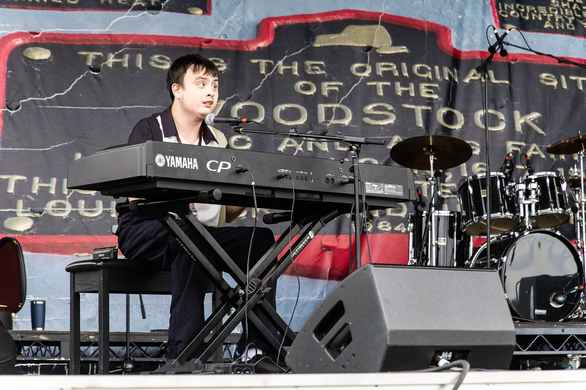 Musician playing Yamaha keyboard on stage with black drum set.
