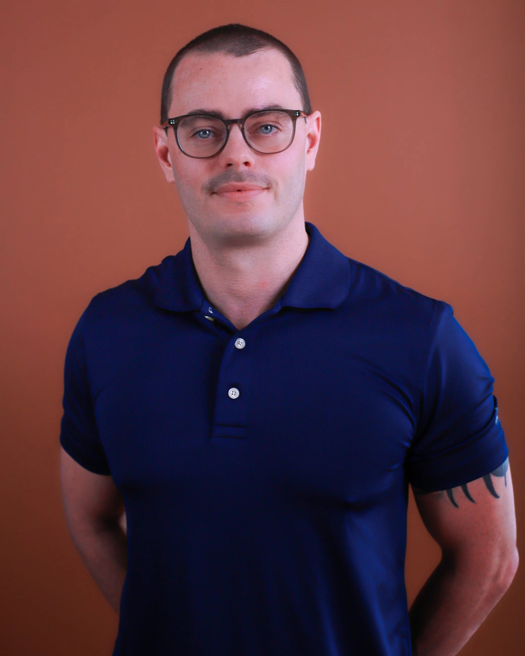 Portrait of a young man with glasses, short hair, wearing a navy blue polo shirt, standing against a brown background.