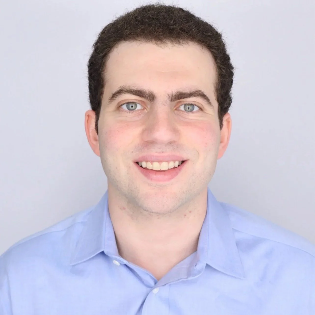 A young man with short dark curly hair and blue eyes, smiling, wearing a light blue collared shirt against a light gray background.