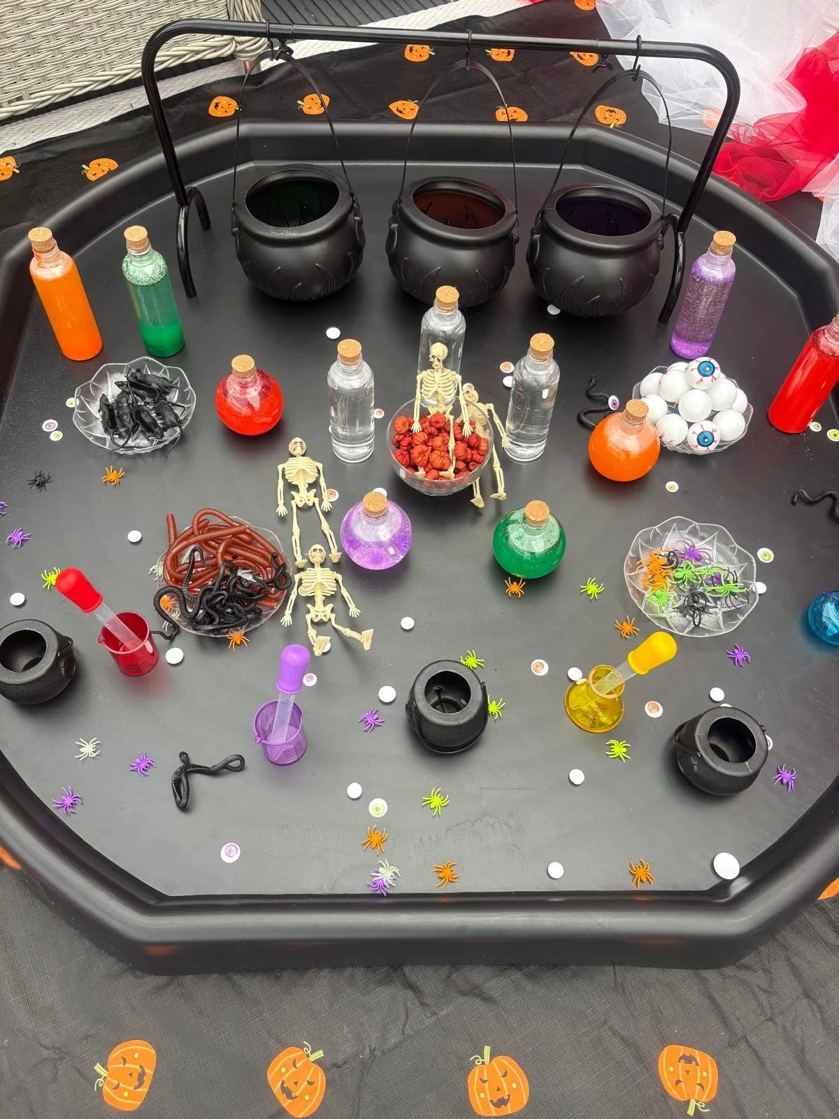 A Halloween-themed game setup with black cauldrons, colorful bottles, plastic skeletons, worms, eyeballs, small spiders, and confetti on a black tray and tablecloth decorated with pumpkin images.