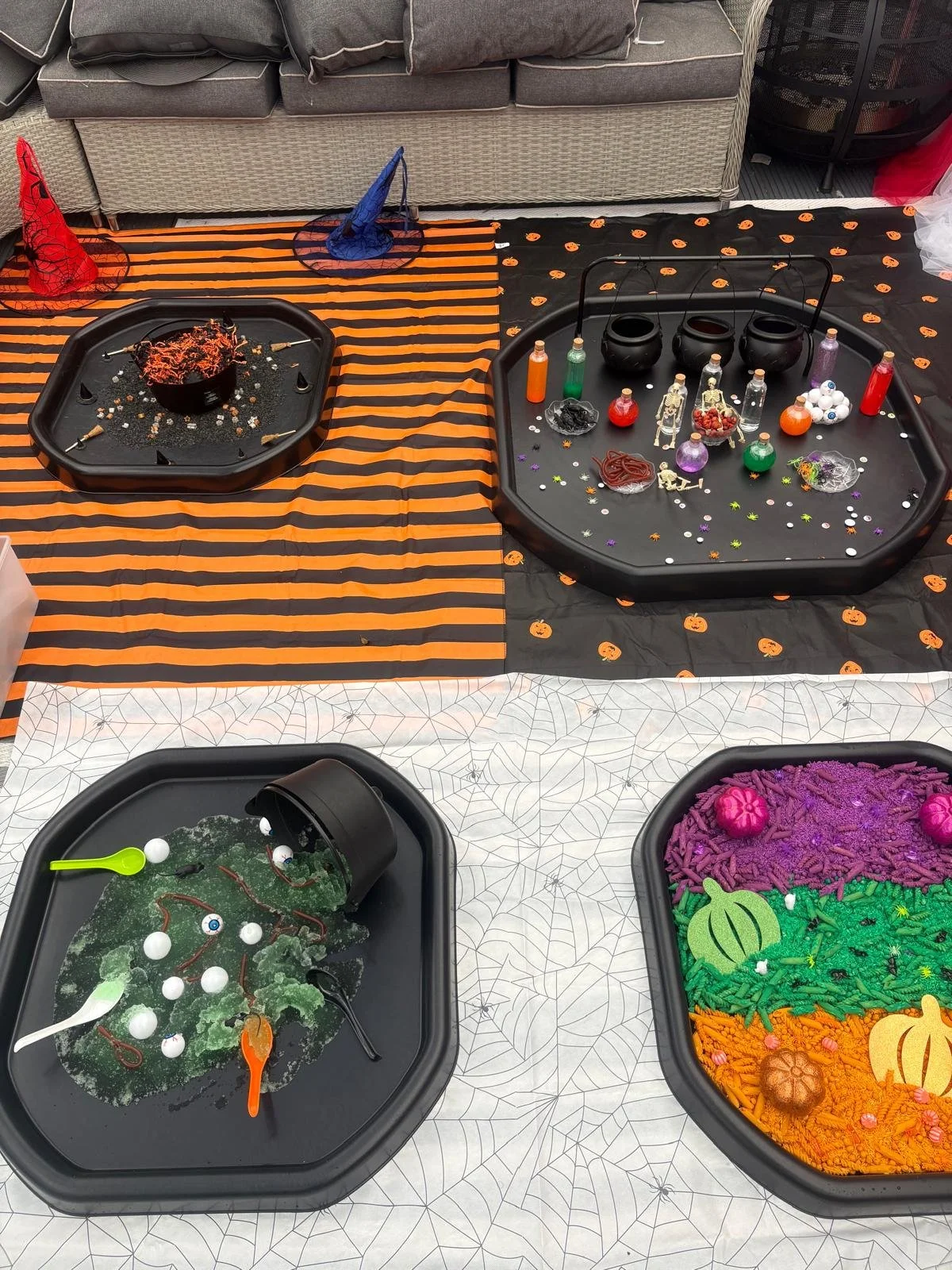 Halloween-themed craft tables with decorations, slime, and small spooky accessories, set on striped orange and black, and white tablecloths with spiderweb patterns.