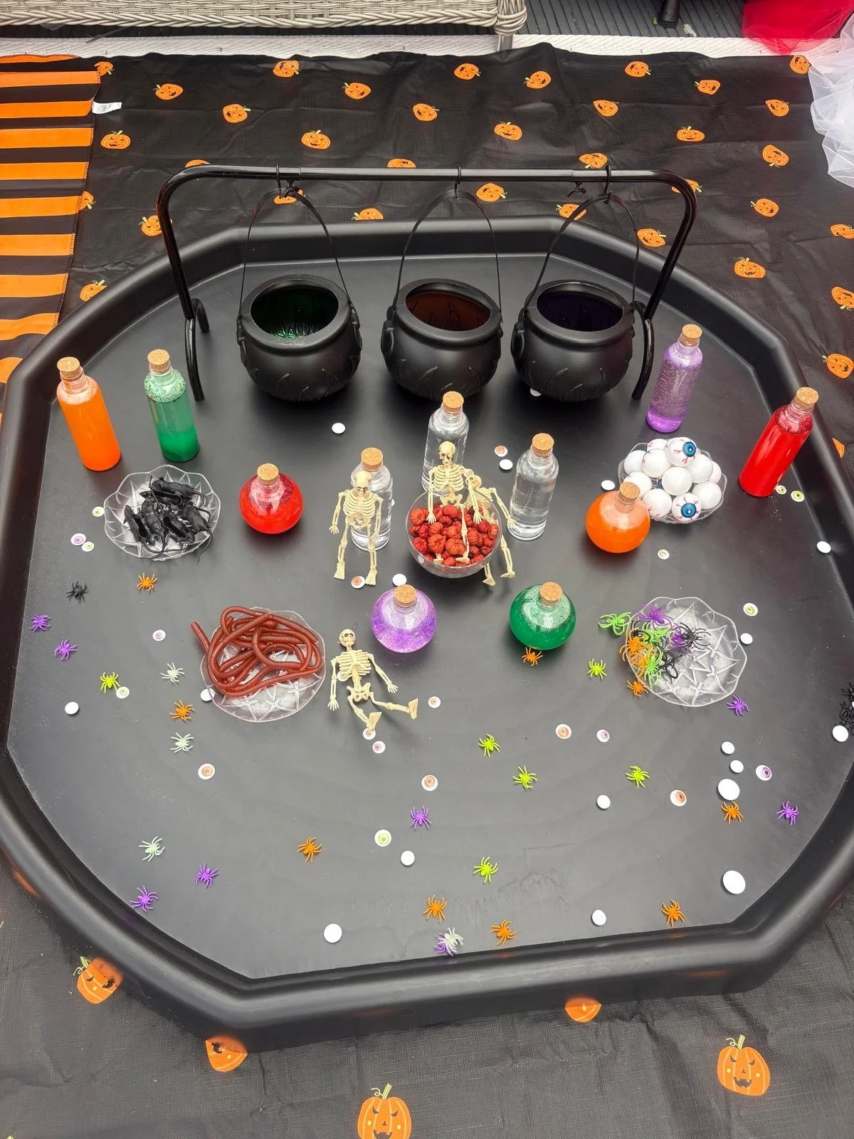 Halloween potion play setup with mini cauldrons, coloured liquids, skeletons, plastic spiders, eyeballs and spooky decorations on a pumpkin tablecloth.