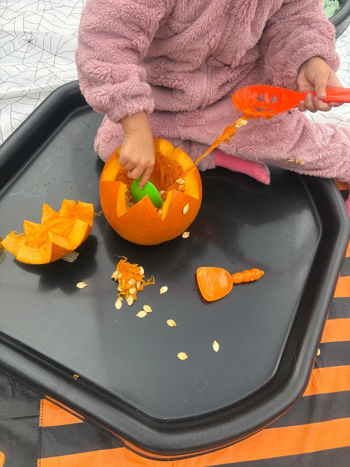 Child in pink fuzzy clothing scooping out pumpkin guts with orange Halloween-themed tools on black tray.