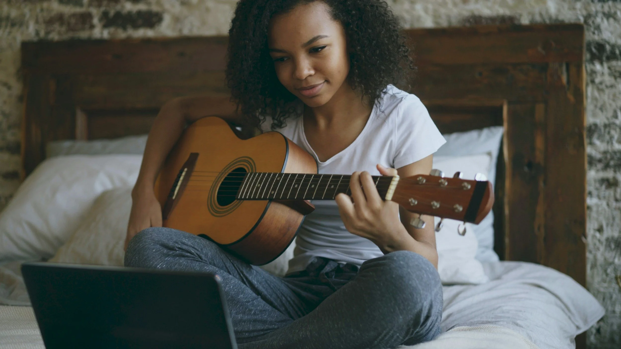 A young woman with curly hair, wearing a white t-shirt and gray pants, sitting on a bed and playing an acoustic guitar. There is a laptop in front of her and a laptop on her lap. The background features a stone wall and a wooden headboard.