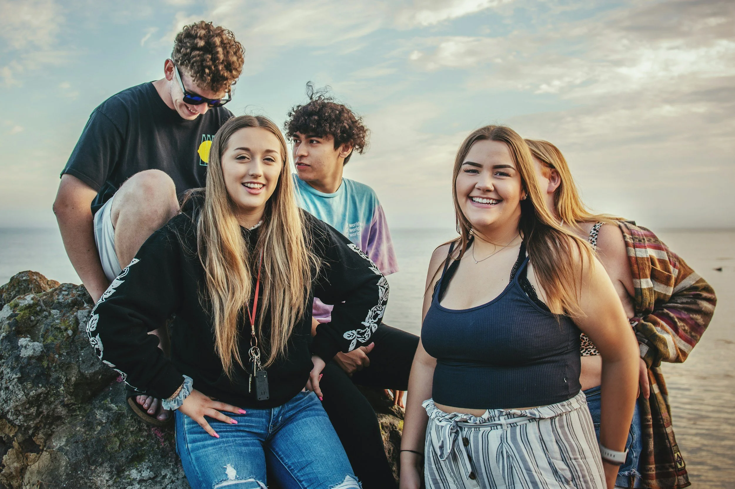 Group of five young friends standing on rocks near the ocean during sunset, smiling and chatting.