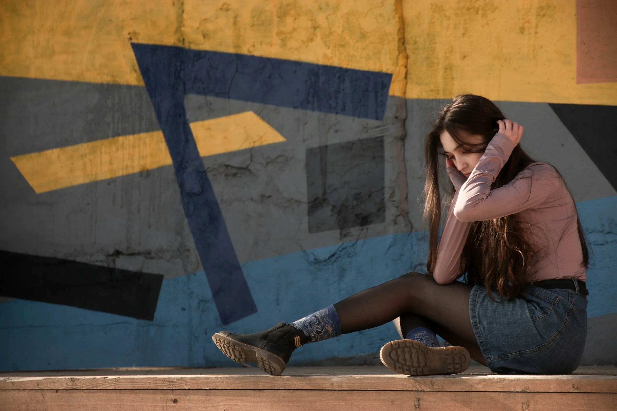 A young woman with long brown hair sitting against a colorful graffiti wall, resting her head on her hands, wearing a pinkish long-sleeve top, denim shorts, and patterned blue tights.