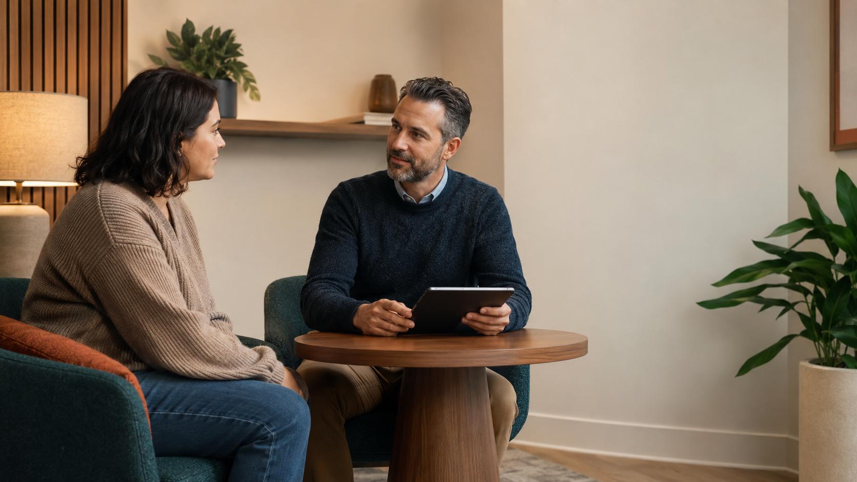 A man and woman sitting in a cozy living room having a conversation, with a tablet in the man's hands.