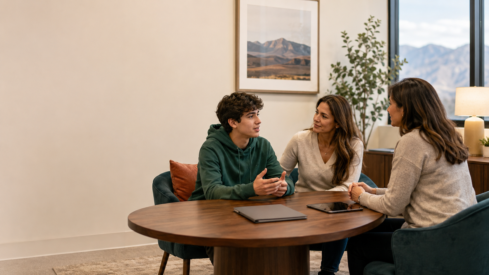 Three people sitting around a wooden table in a cozy room having a face-to-face conversation. One young man and two women are engaged in a discussion. The background shows a large window, a framed landscape picture, a tall potted plant, and a lamp.
